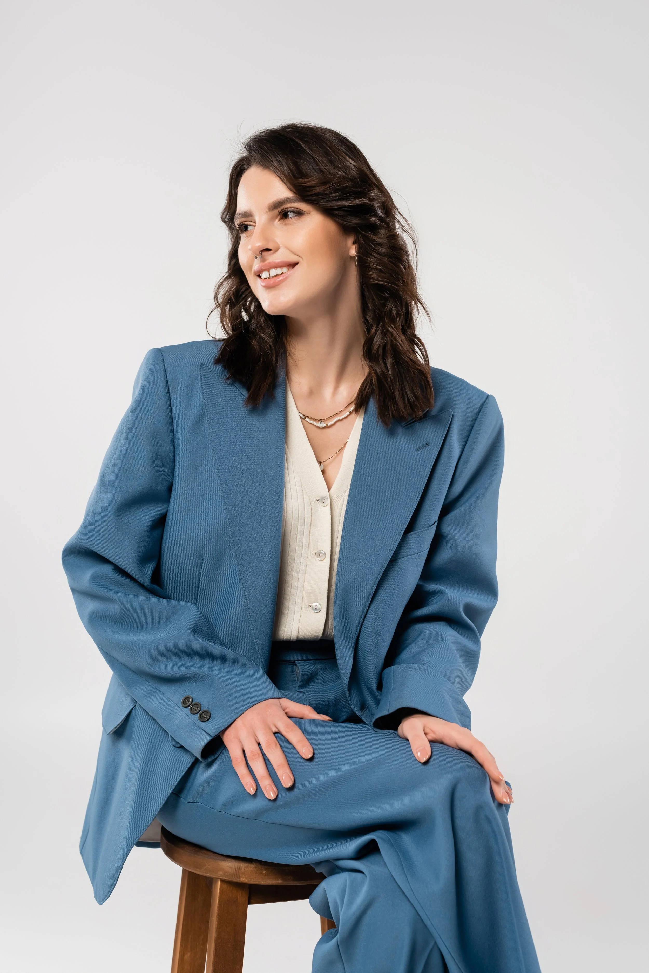 Mujer con cabello castaño oscuro y peinado en ondas, viste un traje azul y una blusa blanca, está sentada en una silla de madera y sonriendo, fondo blanco.