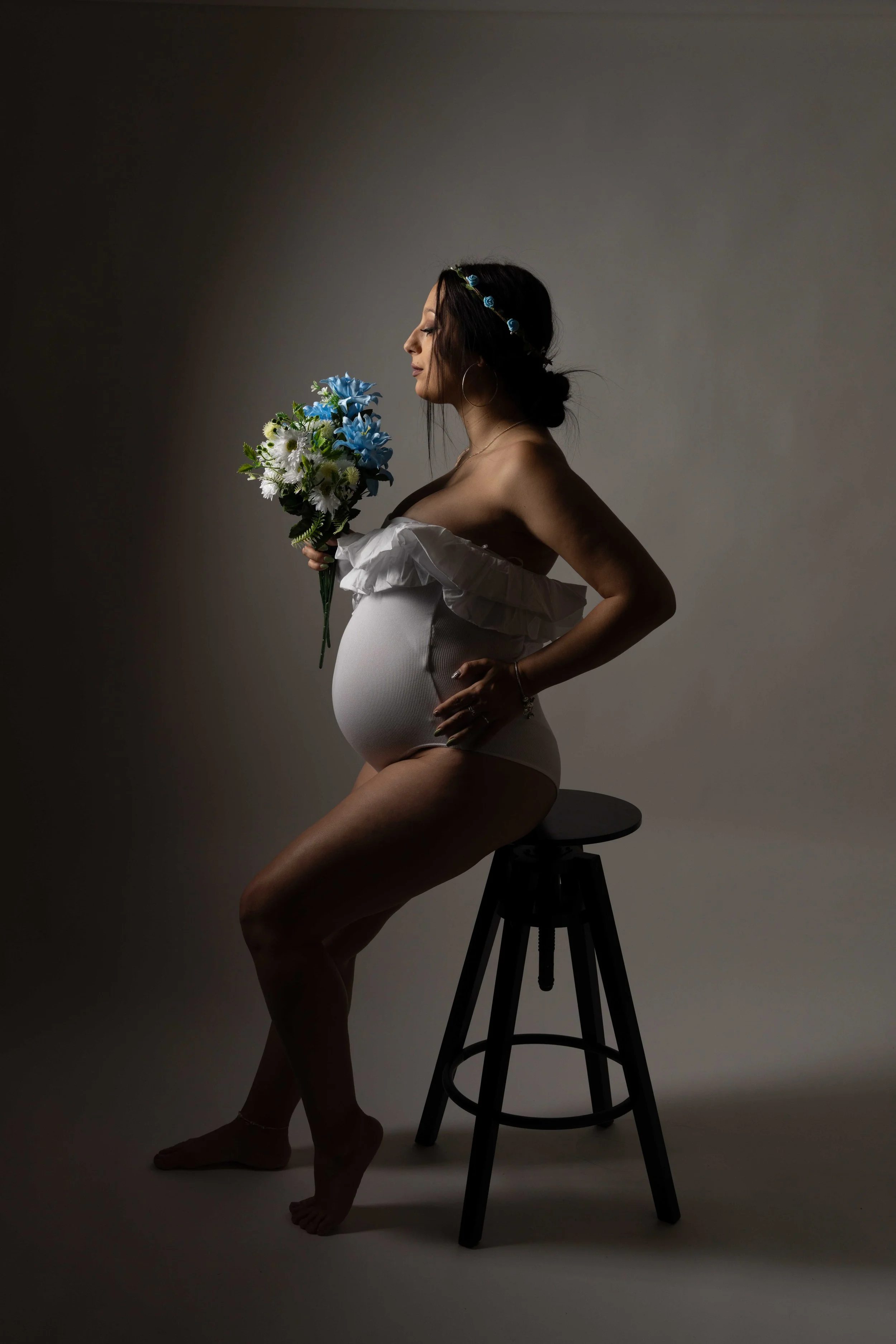Mujer embarazada en una sesión fotográfica, con ropa blanca, sosteniendo un ramo de flores azules y blancas, sentada en un taburete negro, en un fondo neutro, en una pose de perfil con los ojos cerrados.