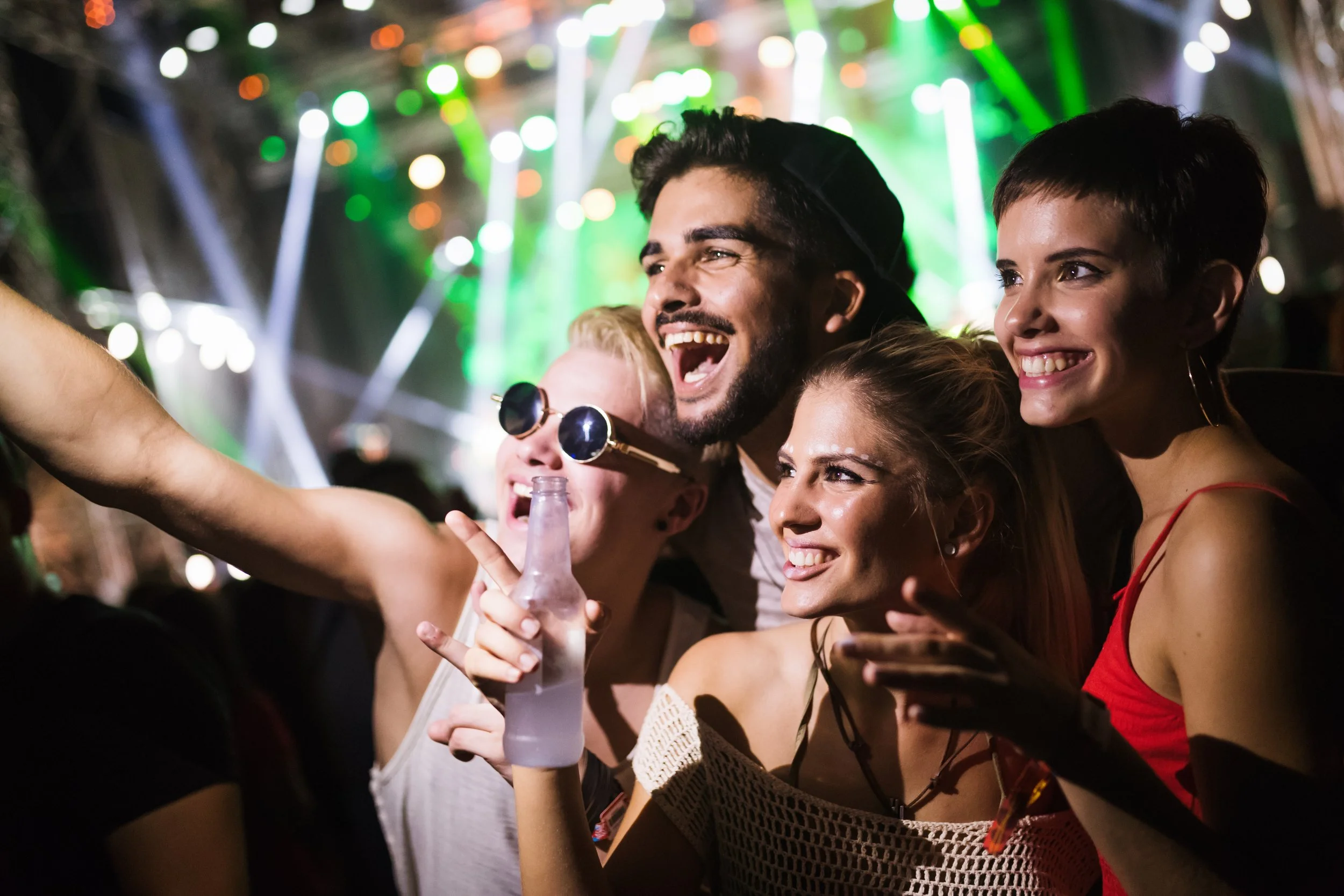 Grupo de amigos disfrutando en un concierto con luces de escenario en el fondo, algunos tomando fotos, riendo y sonriendo.