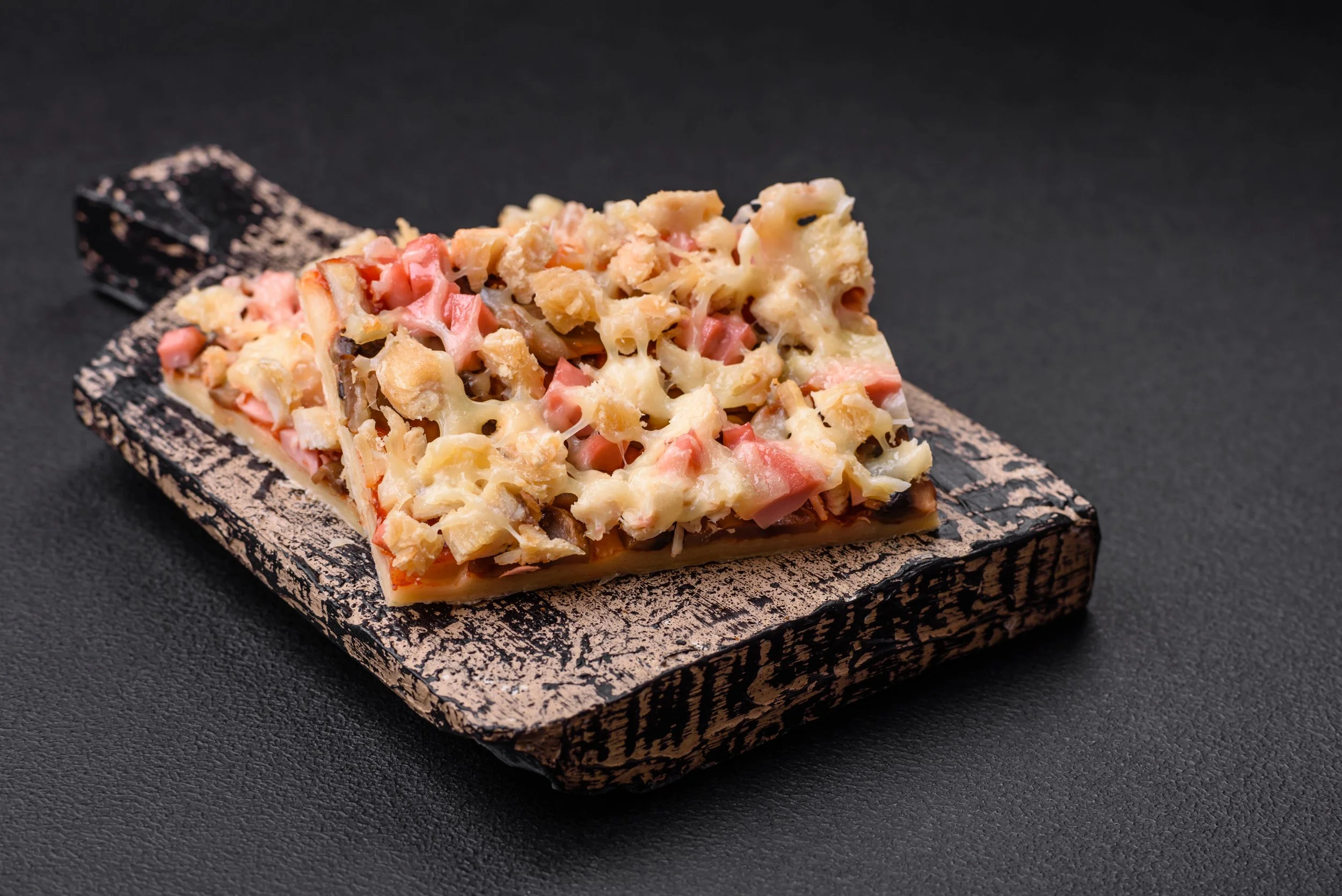 Porción de pizza con queso, jamón, champiñones y otros ingredientes en una tabla de madera sobre fondo negro