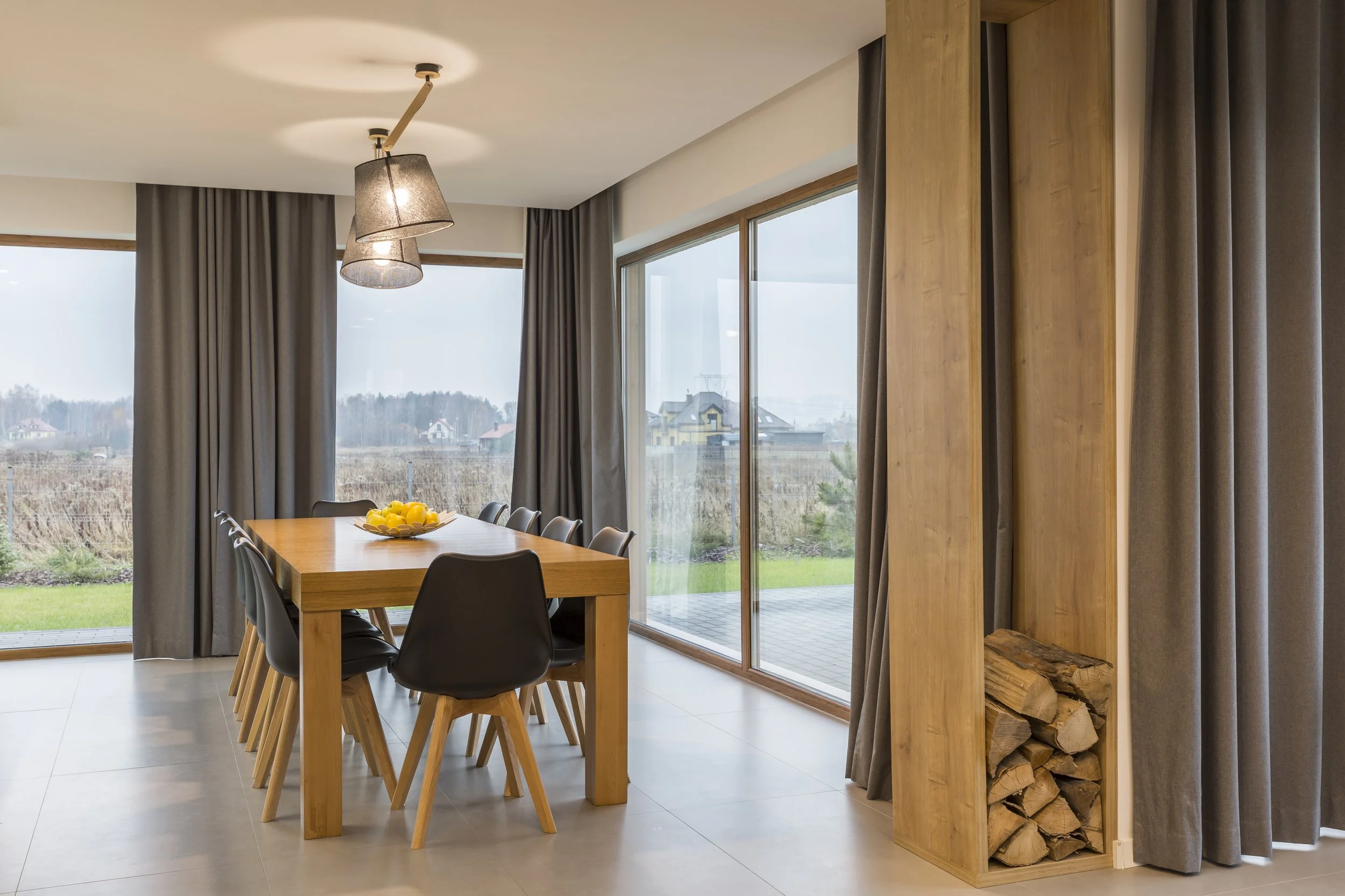 Sala de comedor con mesa de madera y sillas negras, grandes ventanas con cortinas grises y vista a un jardín y campo abierto, estantería empotrada con madera y leña debajo, lámparas de techo de diseño moderno.
