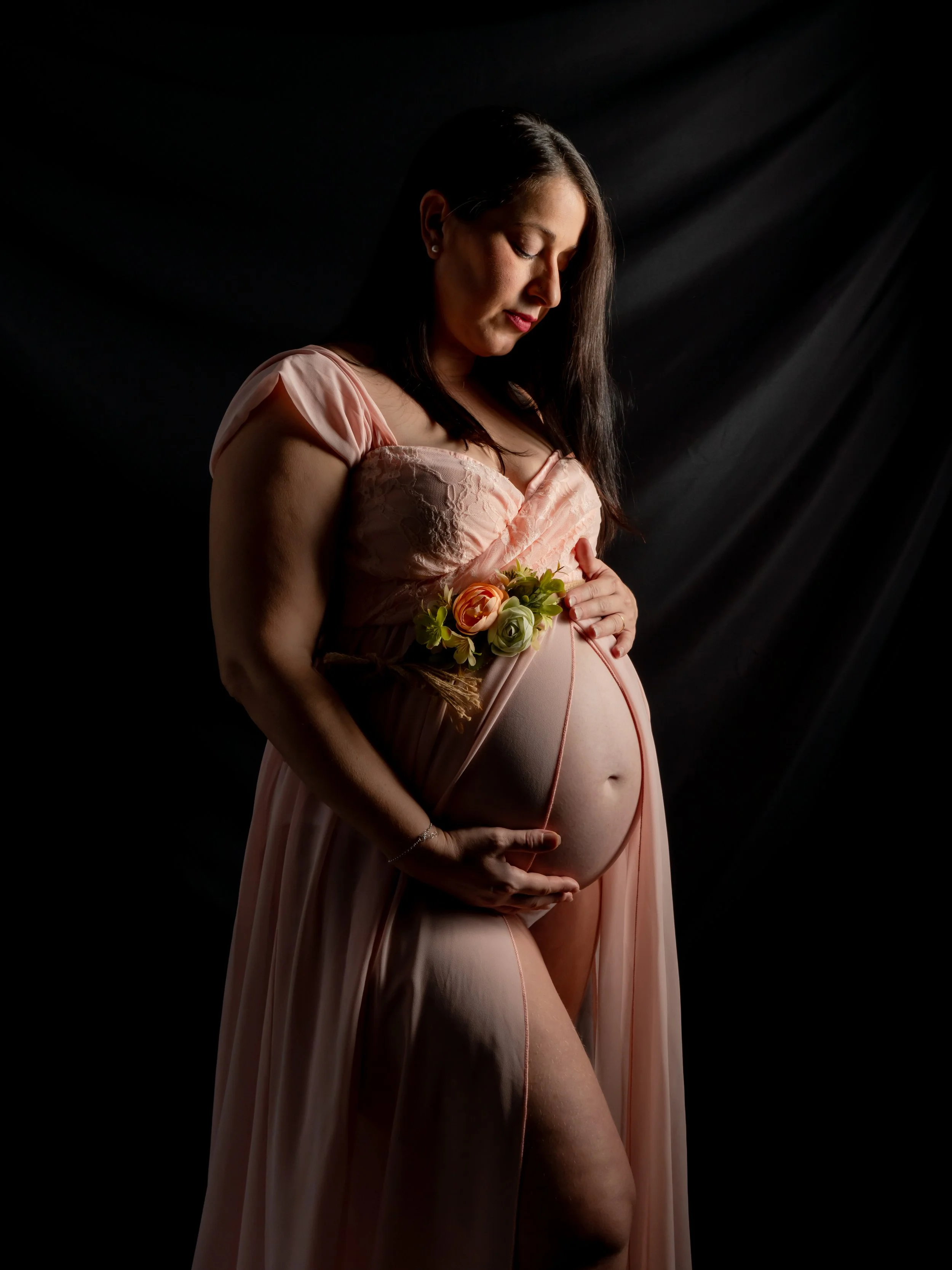 Mujer embarazada con vestido rosa claro sosteniendo su vientre y una flor cerca de su abdomen en un fondo negro.