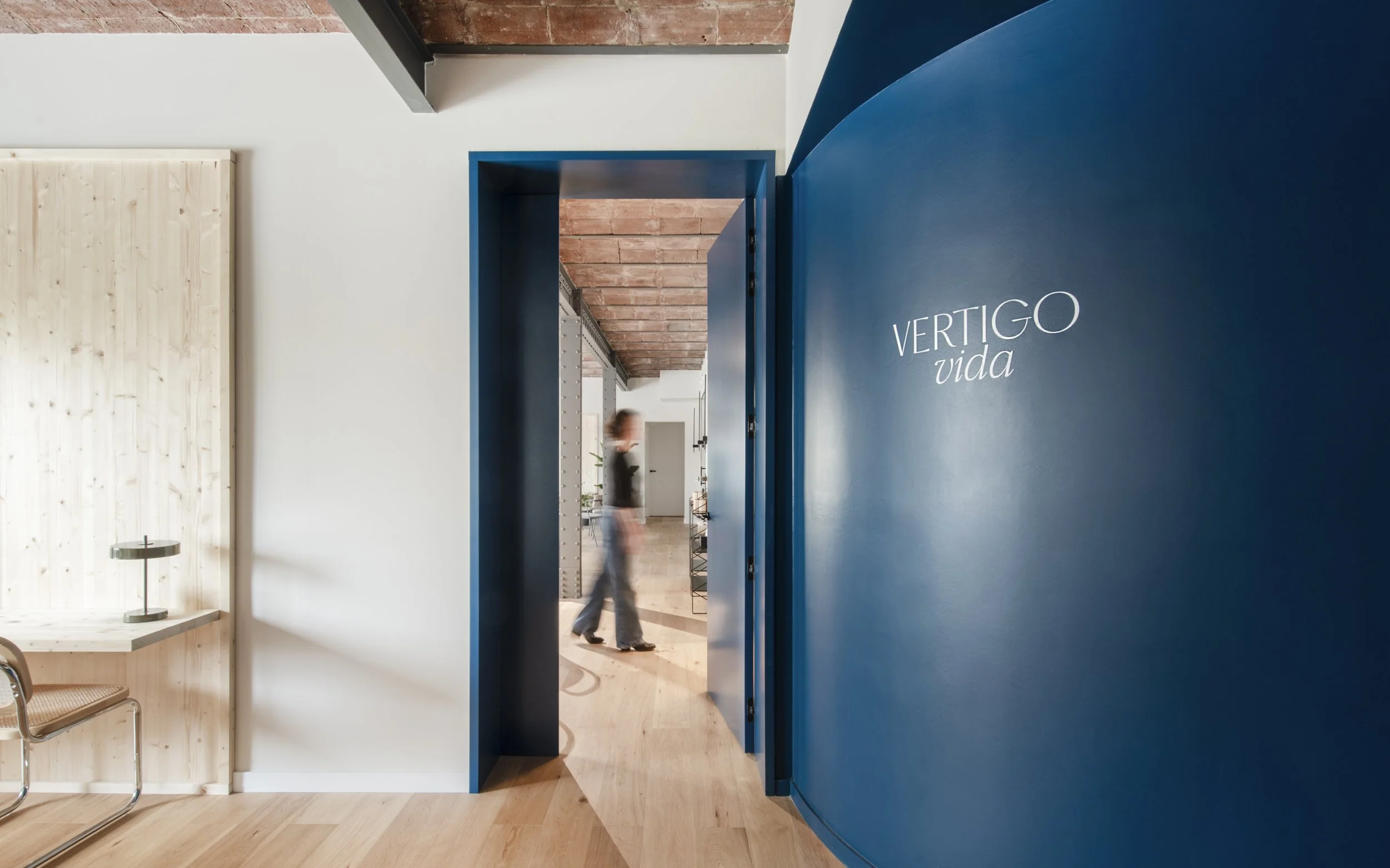 Interior de una oficina moderna con pared azul, puerta abierta y una persona caminando en el fondo.