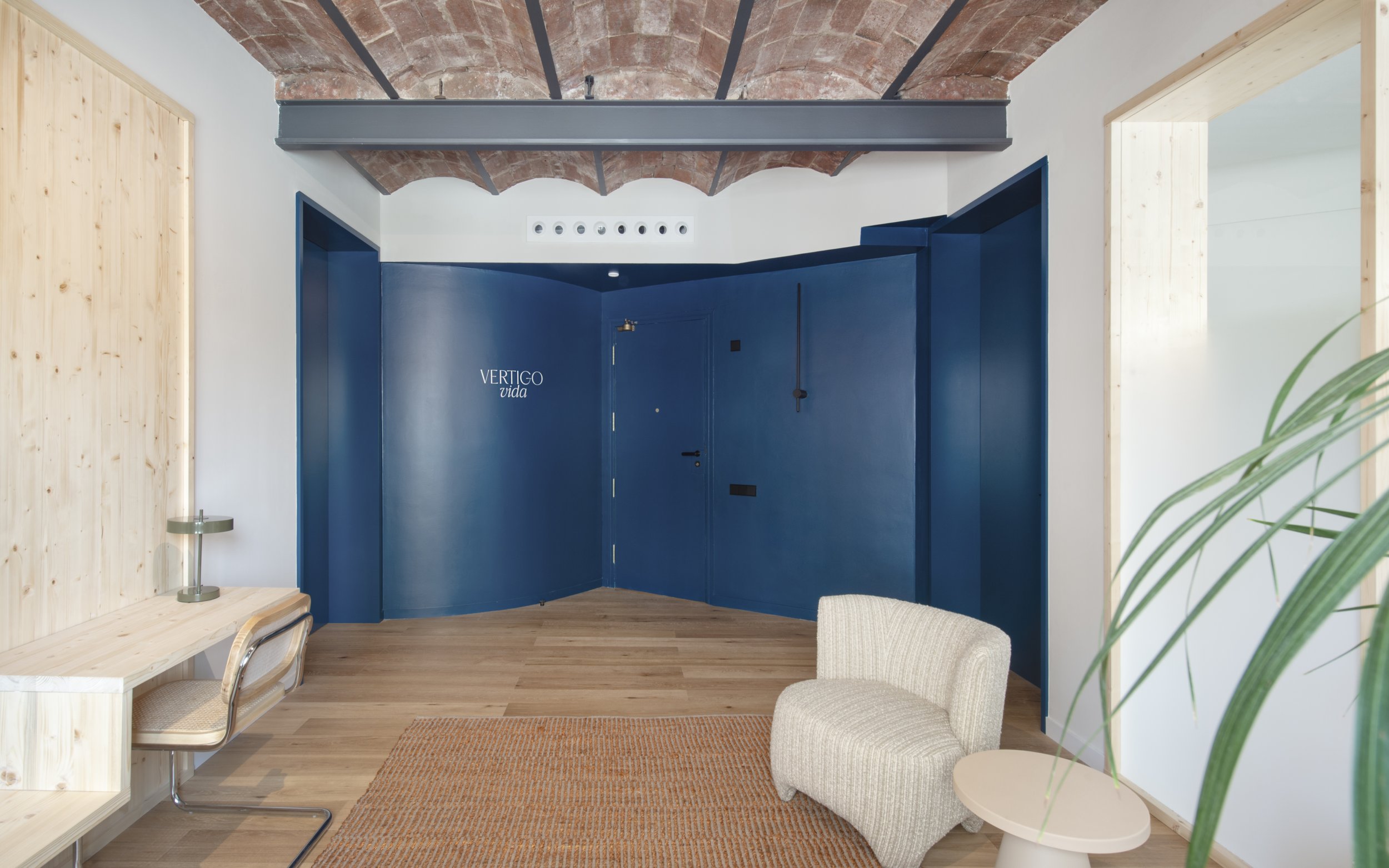 Zona de recepción de Vértigo Vida con pared y puerta en azul petróleo, suelo de madera y techo de bóveda catalana. Interiorismo de Escabeche Studio, Barcelona.
