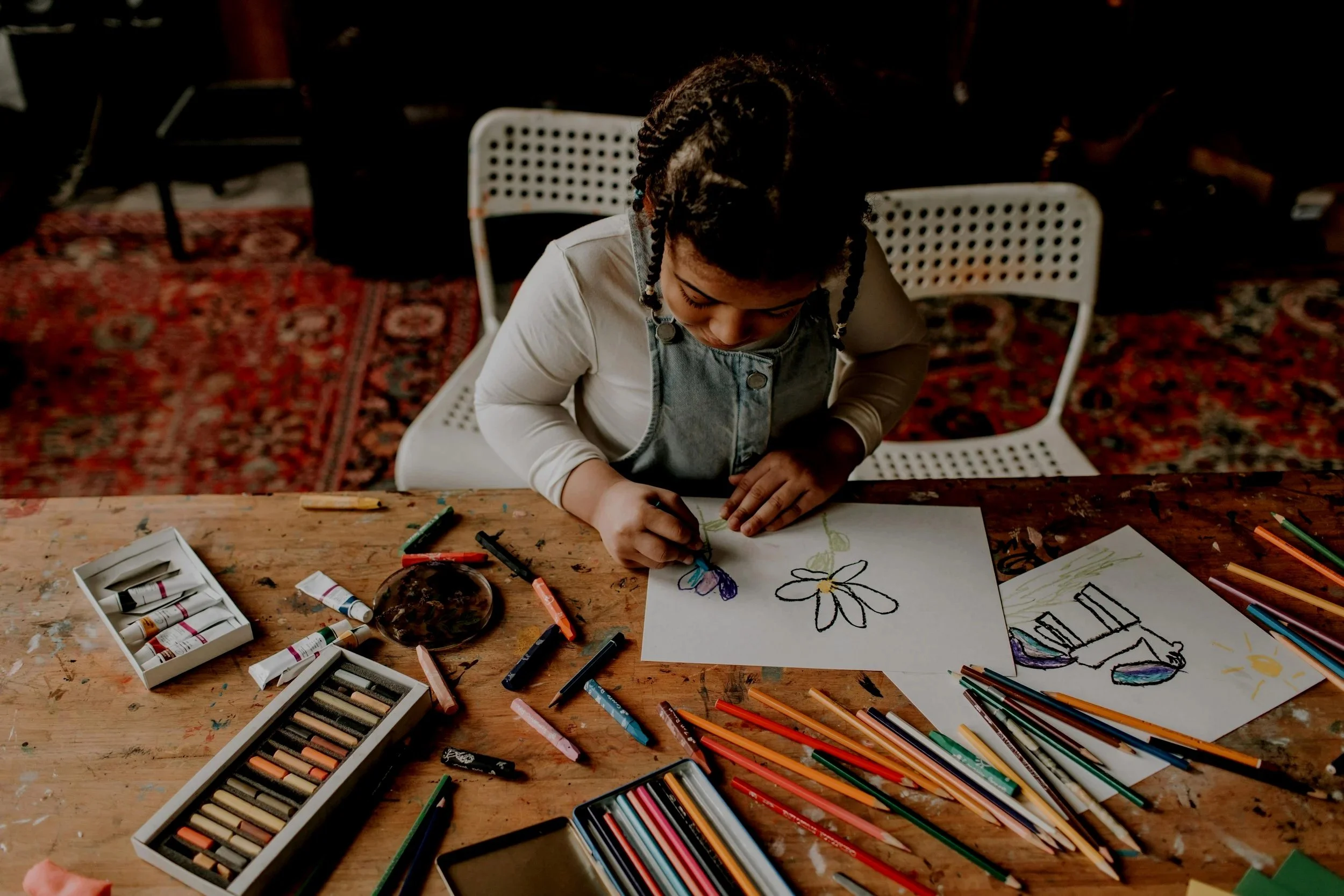 Young girl drawing and coloring at a wooden table with art supplies, including crayons, colored pencils, and drawings of flowers and shoes.
