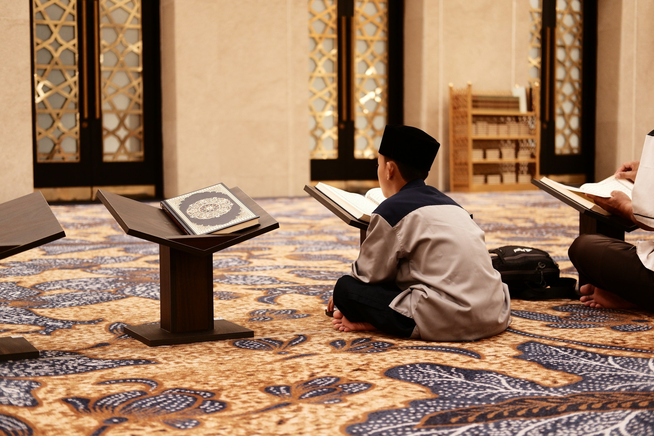 A young boy sitting on a patterned carpet, reading from a religious book on a stand, in a mosque. Other people are also reading nearby.