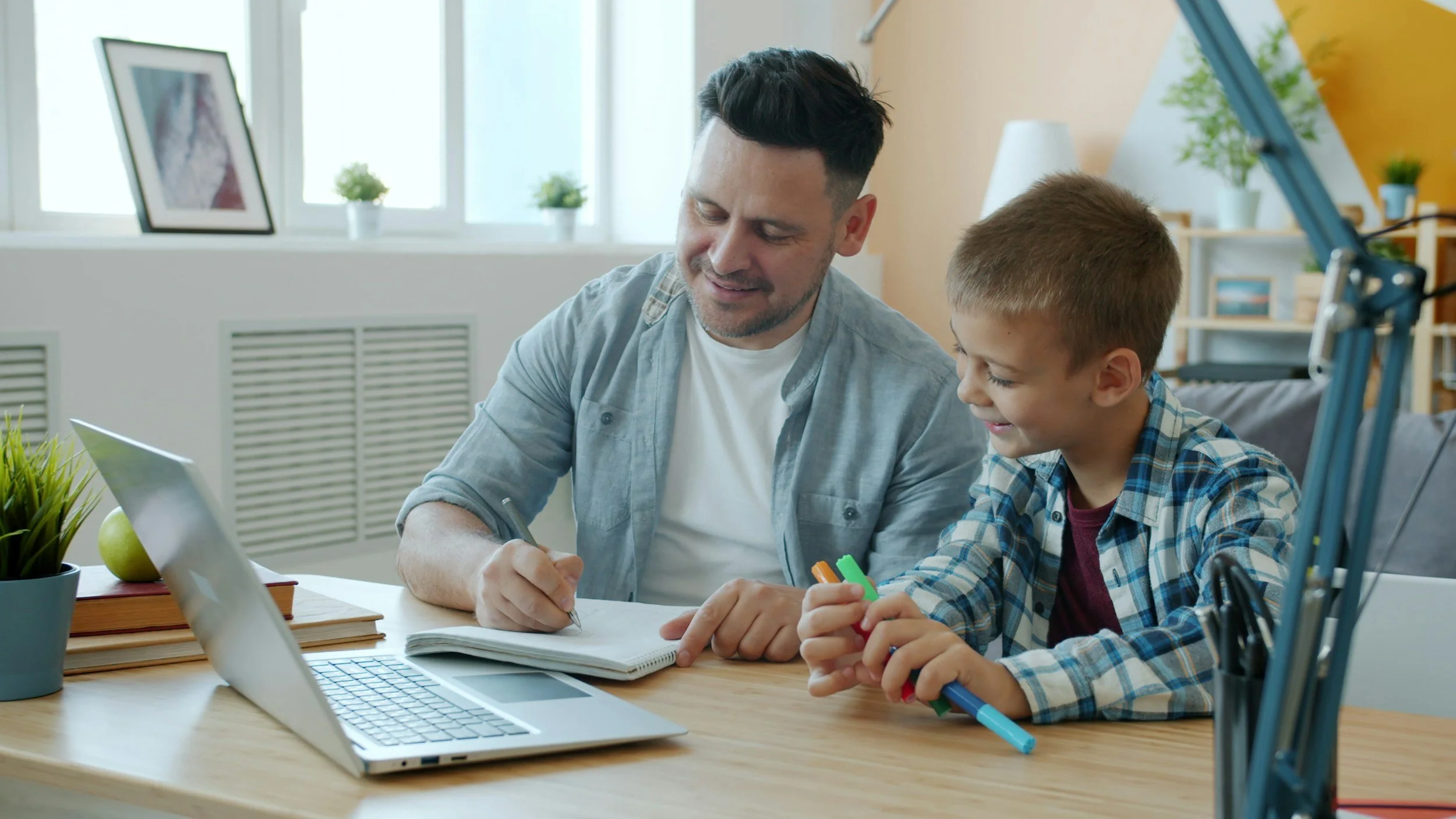 An adult man and a young boy sitting at a wooden desk with a laptop, notebooks, and colorful markers, smiling and engaging in a learning activity in a bright room with large windows and houseplants.
