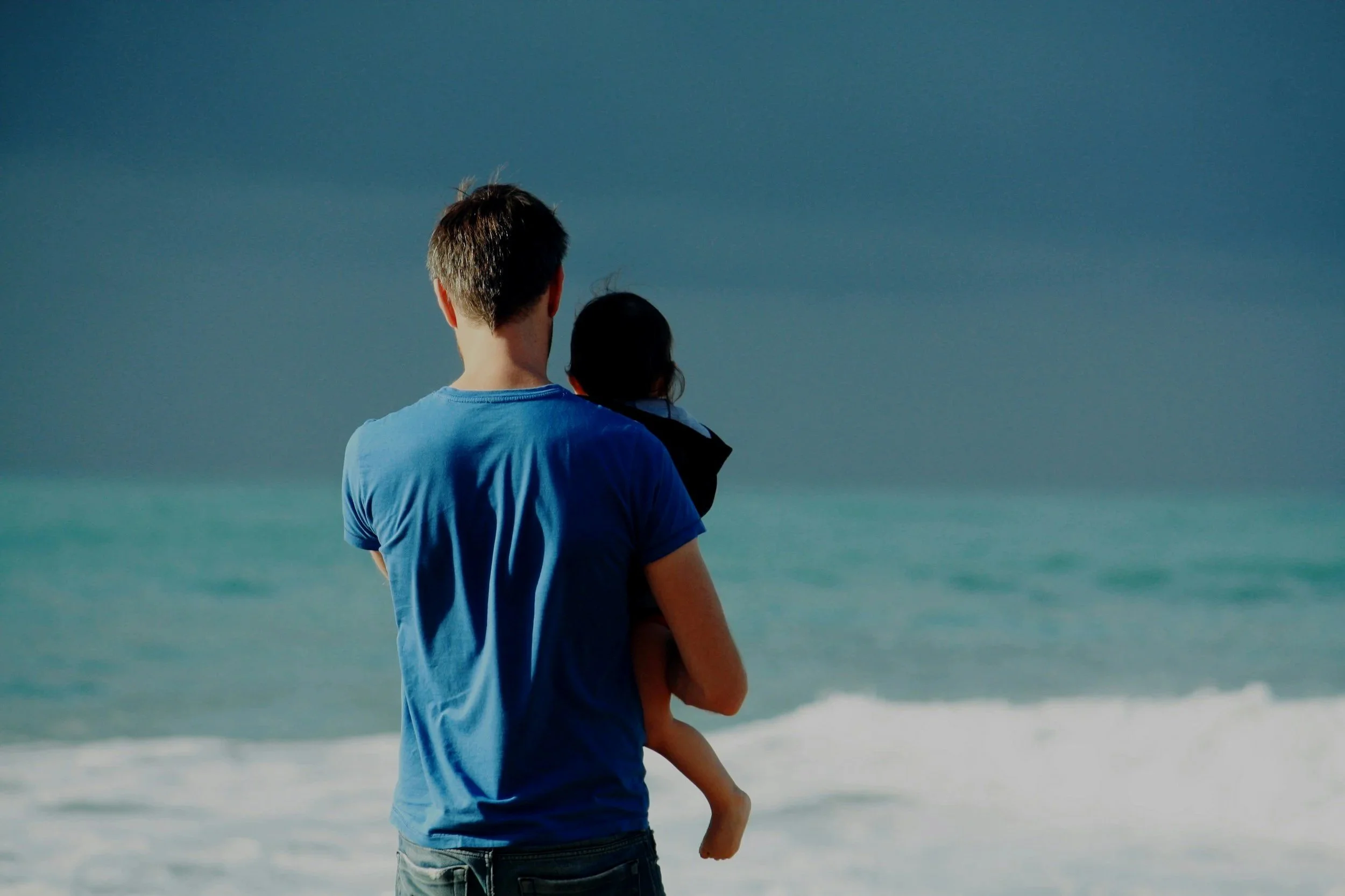 A man holding a child on the beach, facing the ocean with dark clouds in the sky.