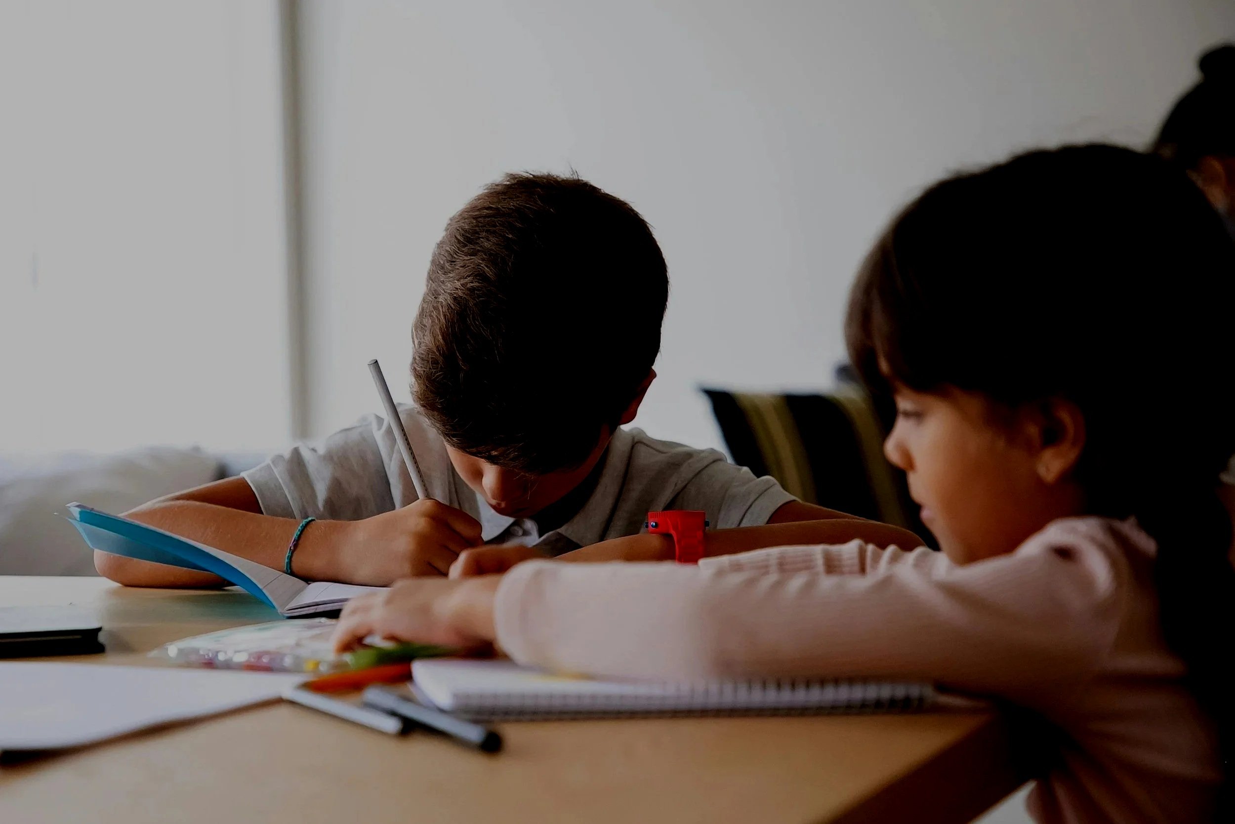 Two children working on homework at a table, surrounded by notebooks, pens, and school supplies.