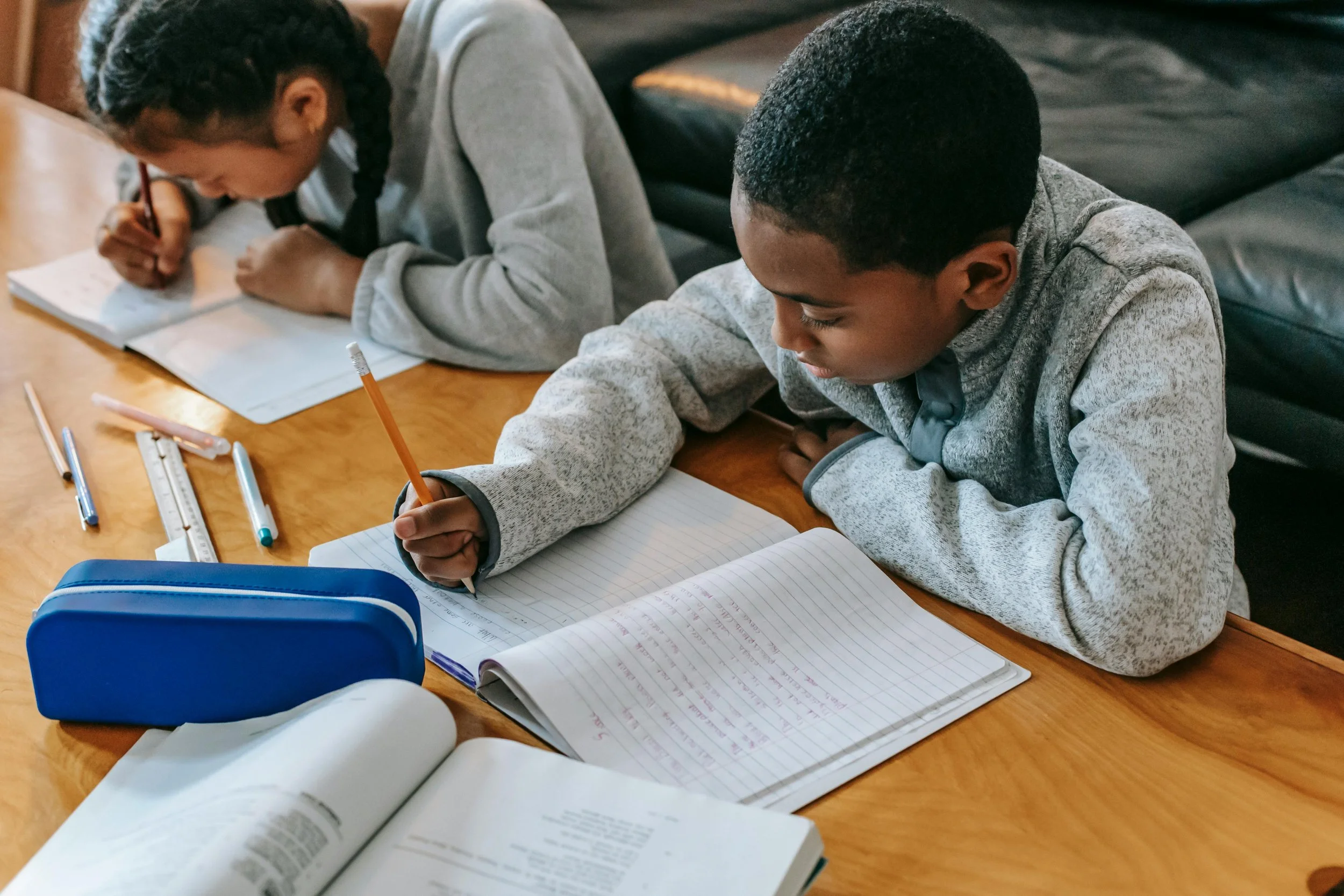 Two children working on homework at a wooden table with books, notebooks, and writing tools.