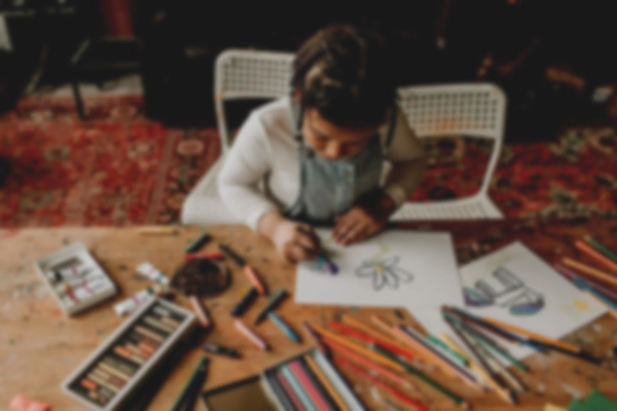 Young girl drawing and coloring at a wooden table with art supplies, including crayons, colored pencils, and drawings of flowers and shoes.