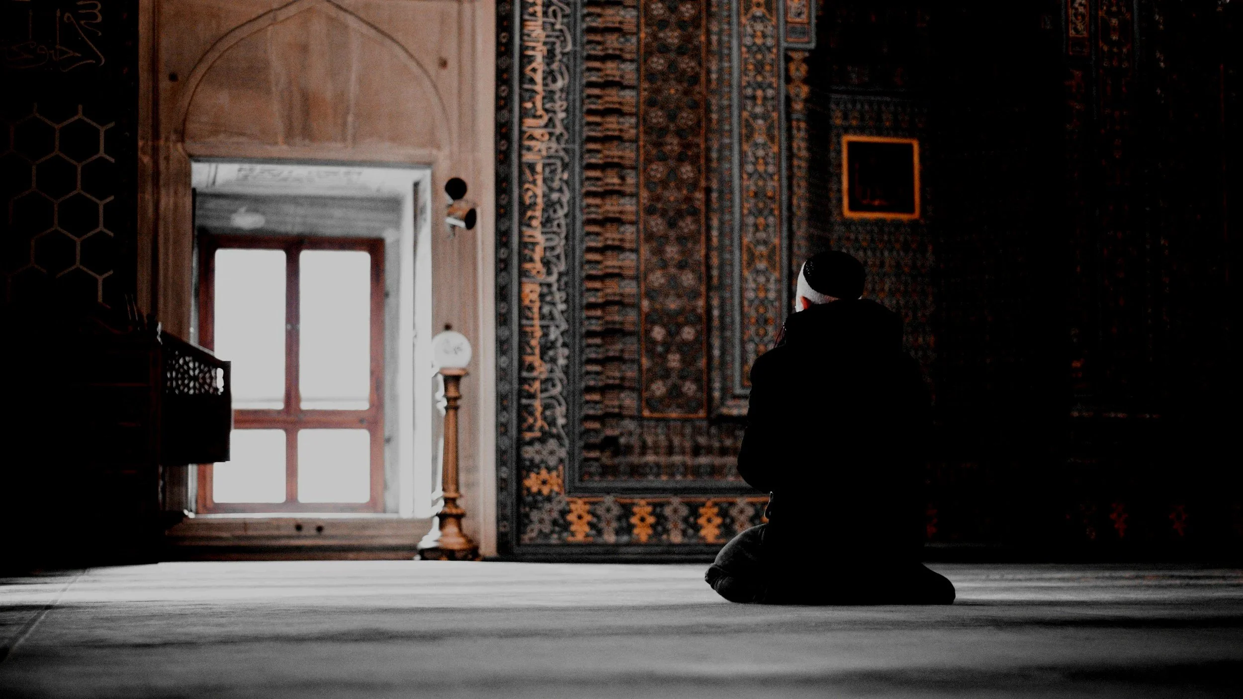 Person kneeling and praying inside a mosque, with an ornate carpet on the wall and a window letting in natural light.