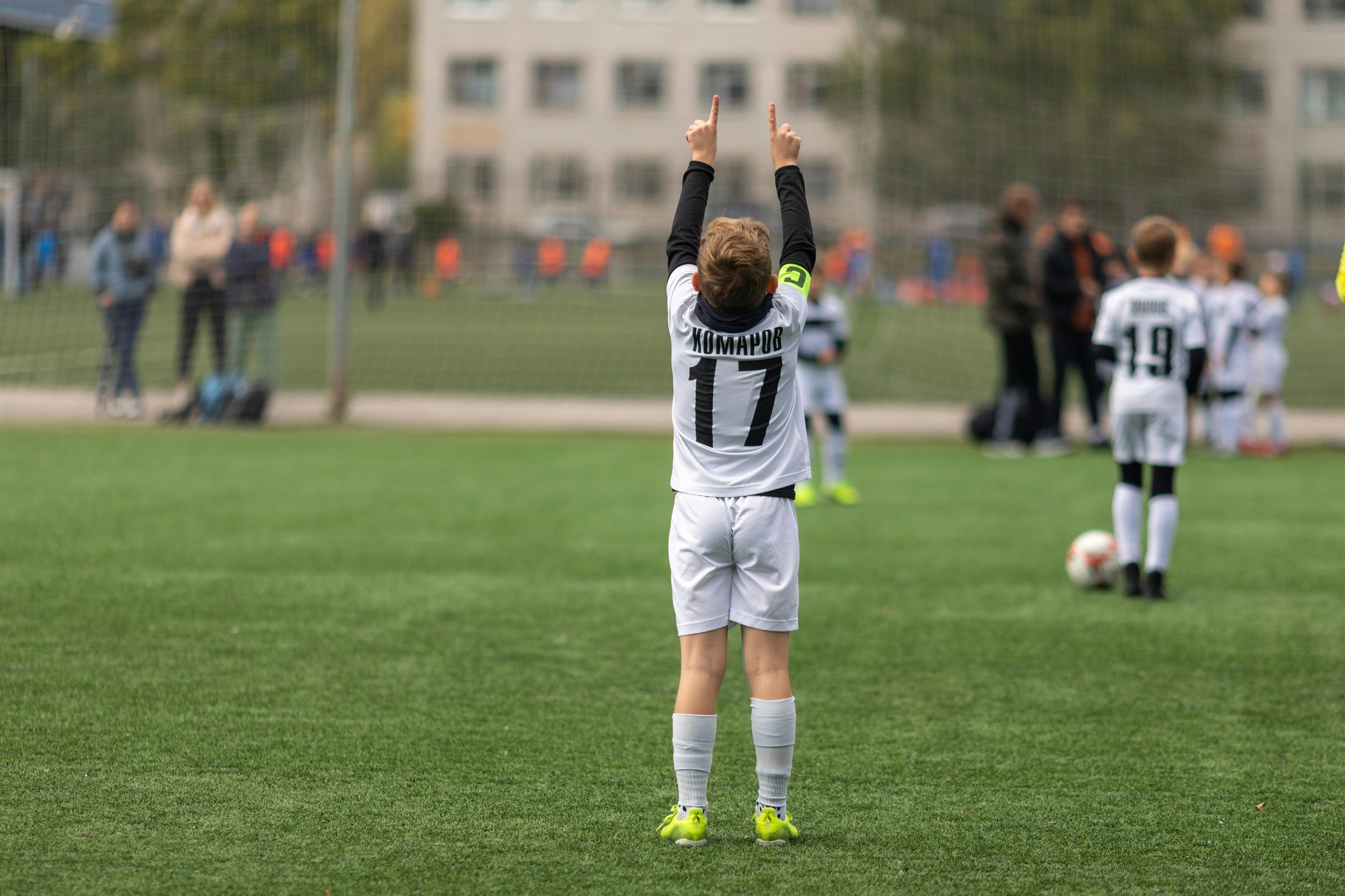 A young soccer player wearing a white jersey with the number 17, standing on a green grass field with arms raised and fingers pointing upward, as other players and spectators are visible in the background.