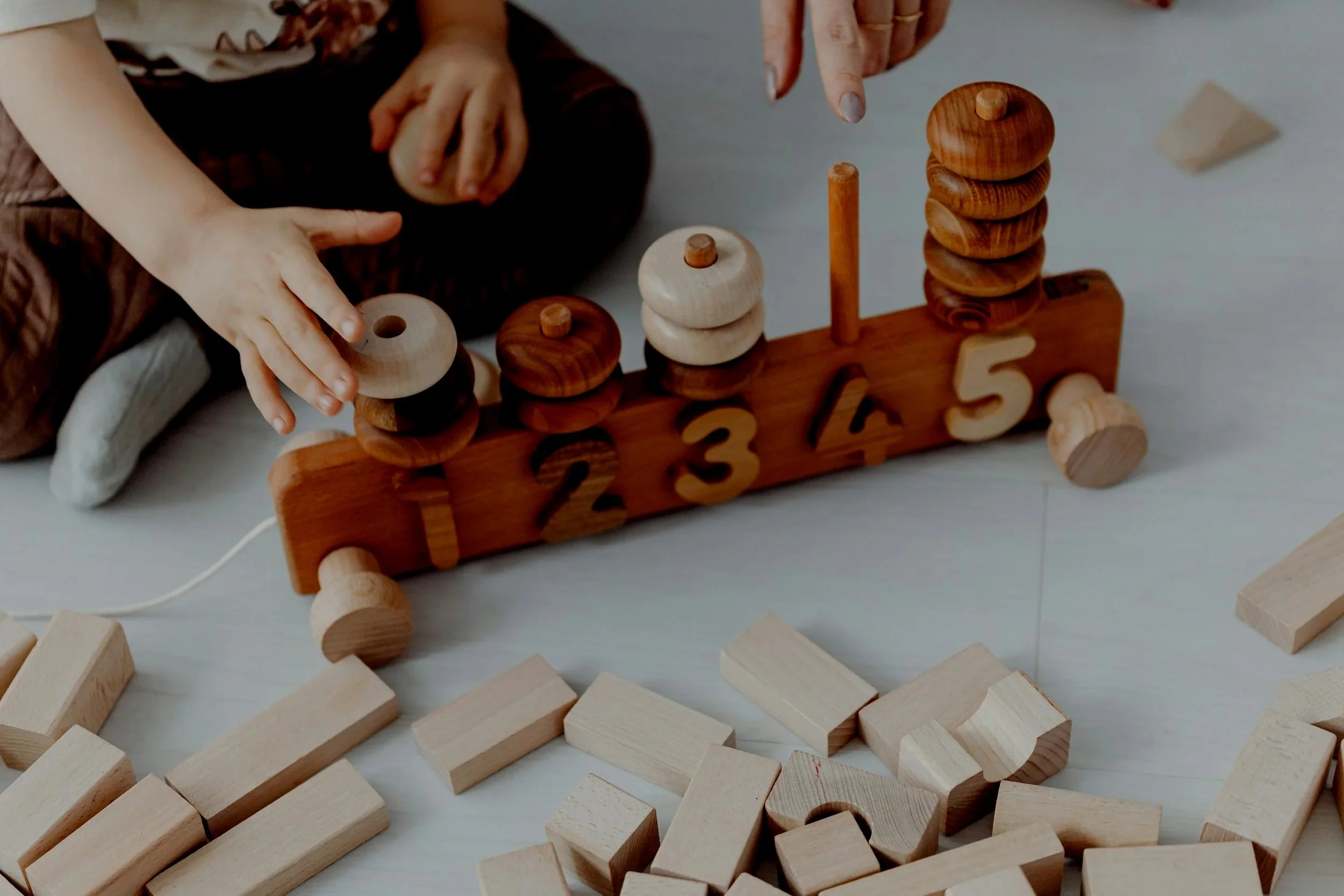 Child and adult playing with a wooden stacking and number toy, with scattered wooden blocks around on the floor.