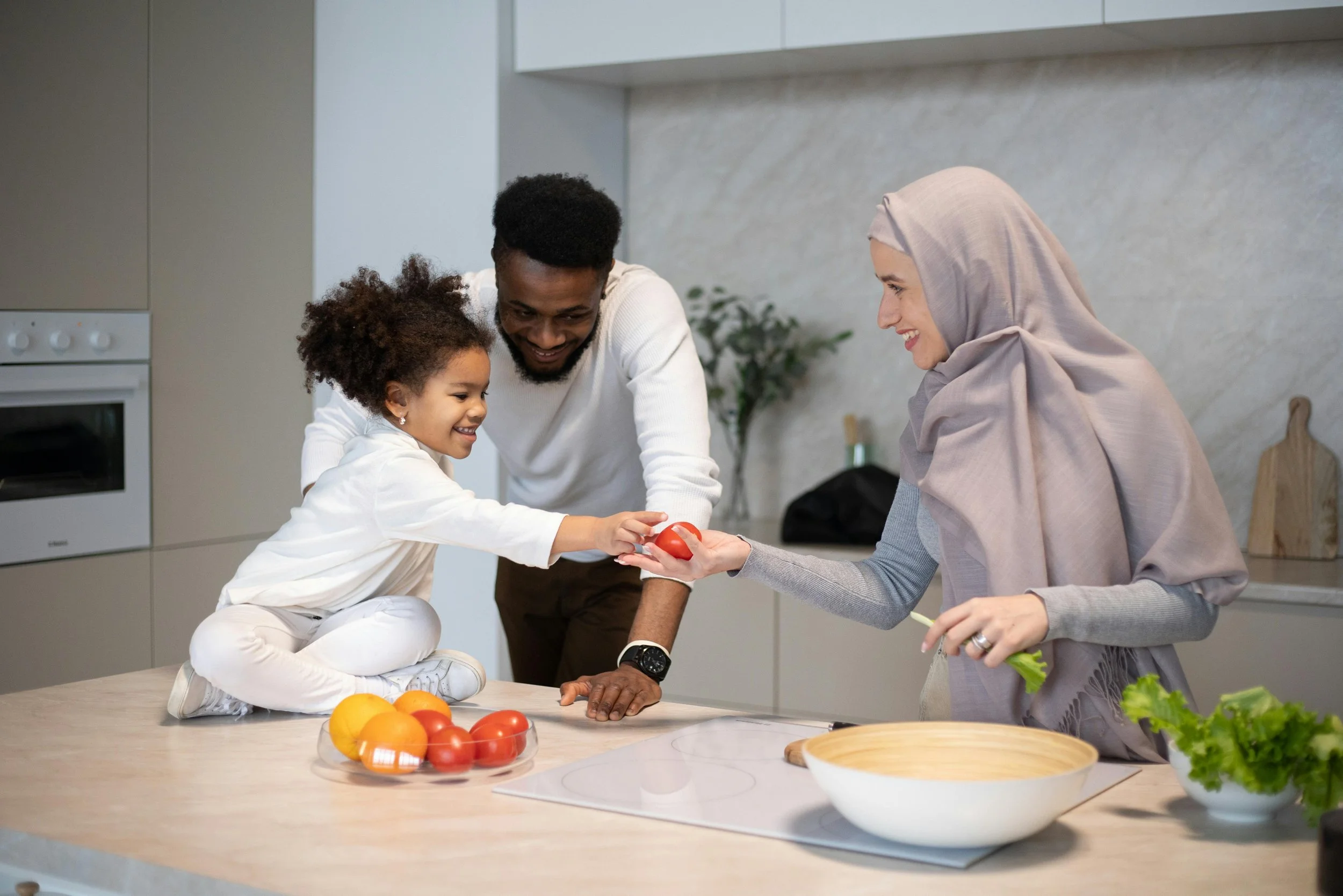 Family enjoying cooking together in a modern kitchen, with a young girl handing a tomato to an adult woman wearing a hijab.