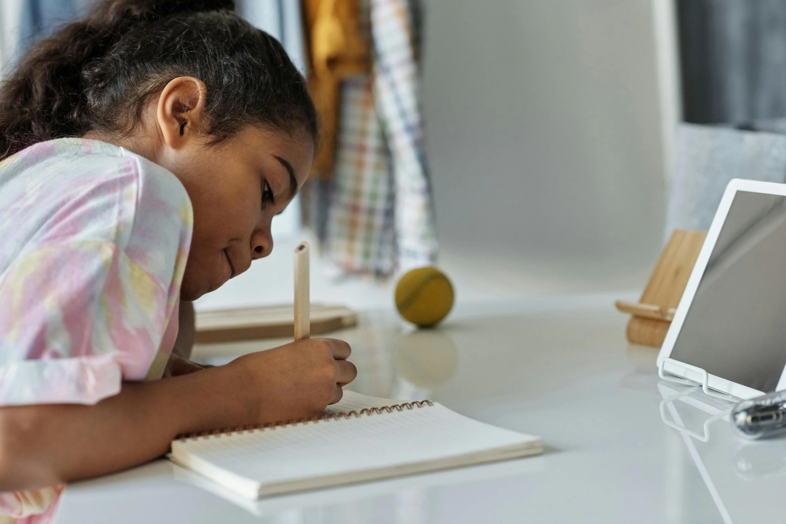 A young girl with dark hair in a ponytail, wearing a pastel tie-dye shirt, is lying on a white table and writing in a notebook with a pencil. There is a tablet, yellow ball, and some wooden objects on the table, with a checkered cloth hanging in the background.