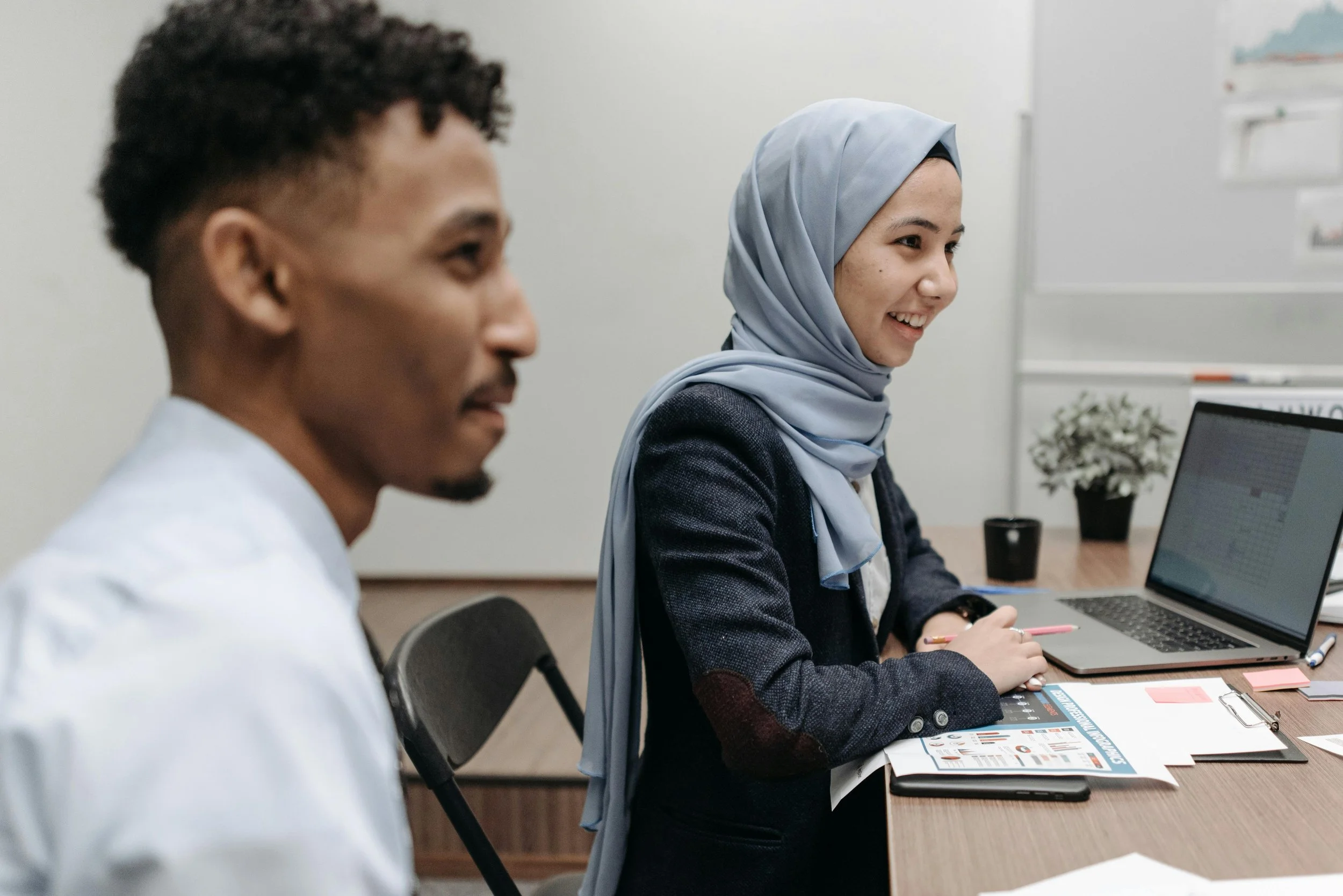 A woman wearing a light blue hijab and a dark blazer sitting at a desk with a laptop, smiling during a meeting or discussion, with a man in the foreground out of focus.
