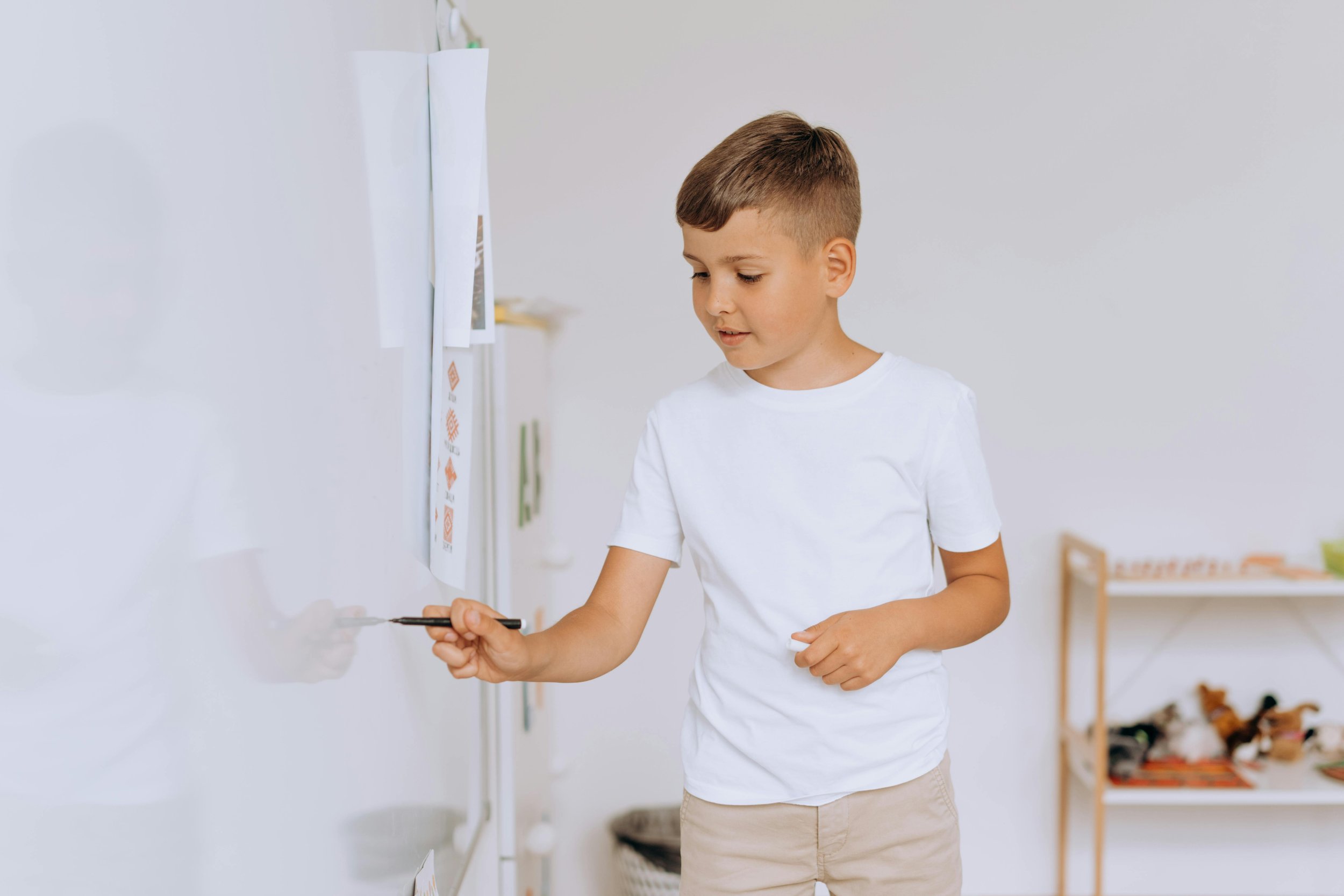 A young boy in a white t-shirt and beige shorts writes on a whiteboard with a black marker in a classroom.