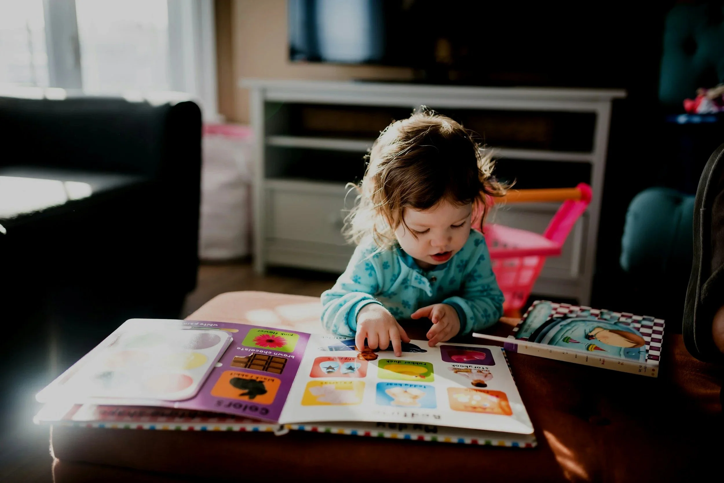 A young girl with curly hair in pajamas sitting at a table, looking at and pointing to picture books with colorful illustrations in a sunlit room.
