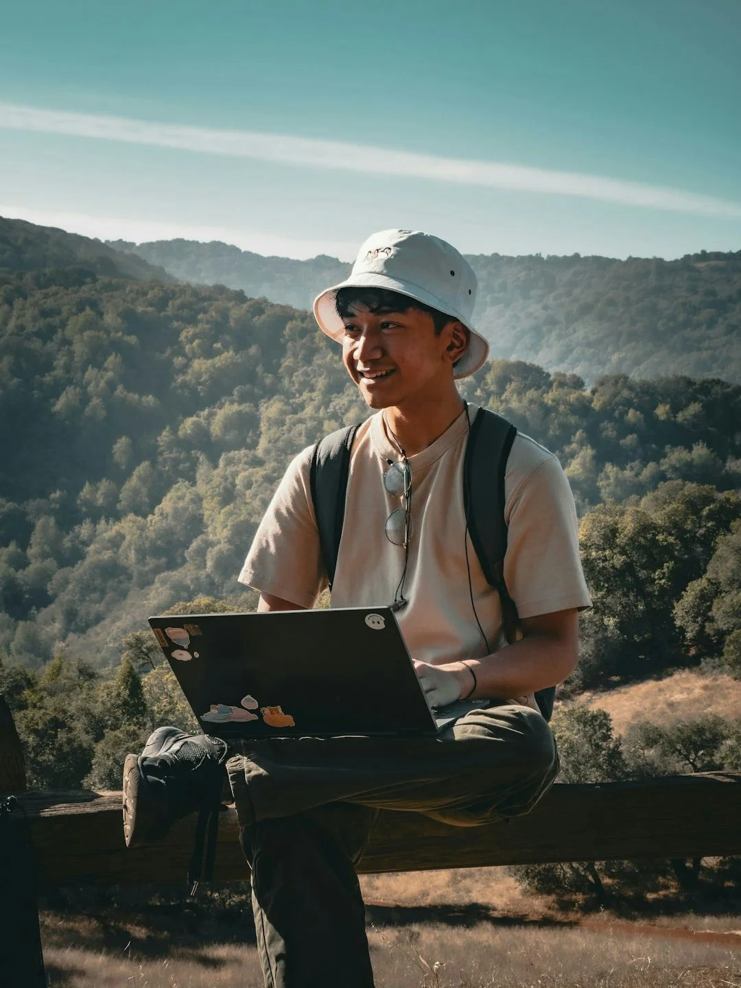 A young man with a white bucket hat, beige T-shirt, and backpack sitting on a wooden log outdoors, working on a laptop with a forested hillside in the background.