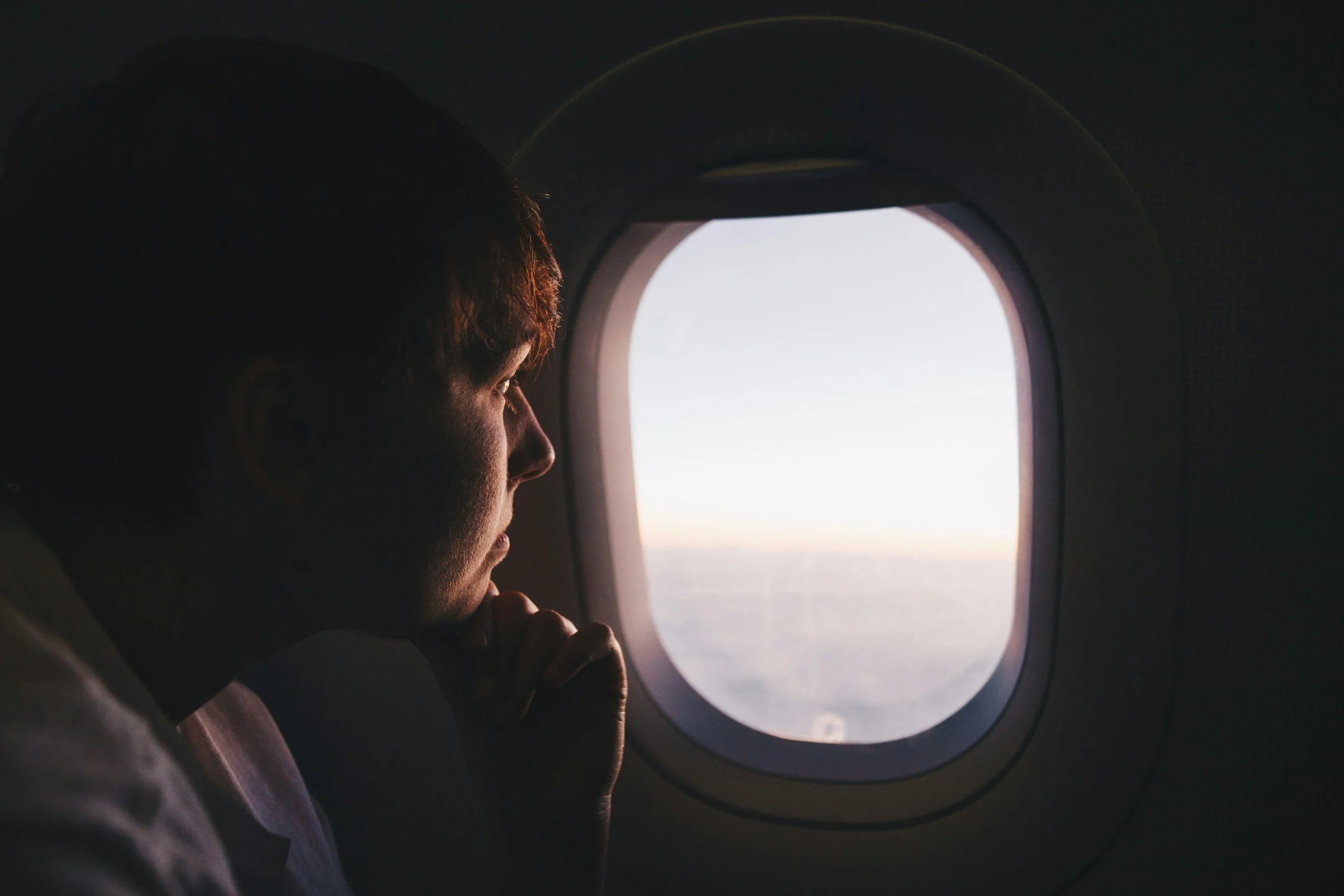 A person with short hair looking out the window of an airplane at sunset or sunrise.