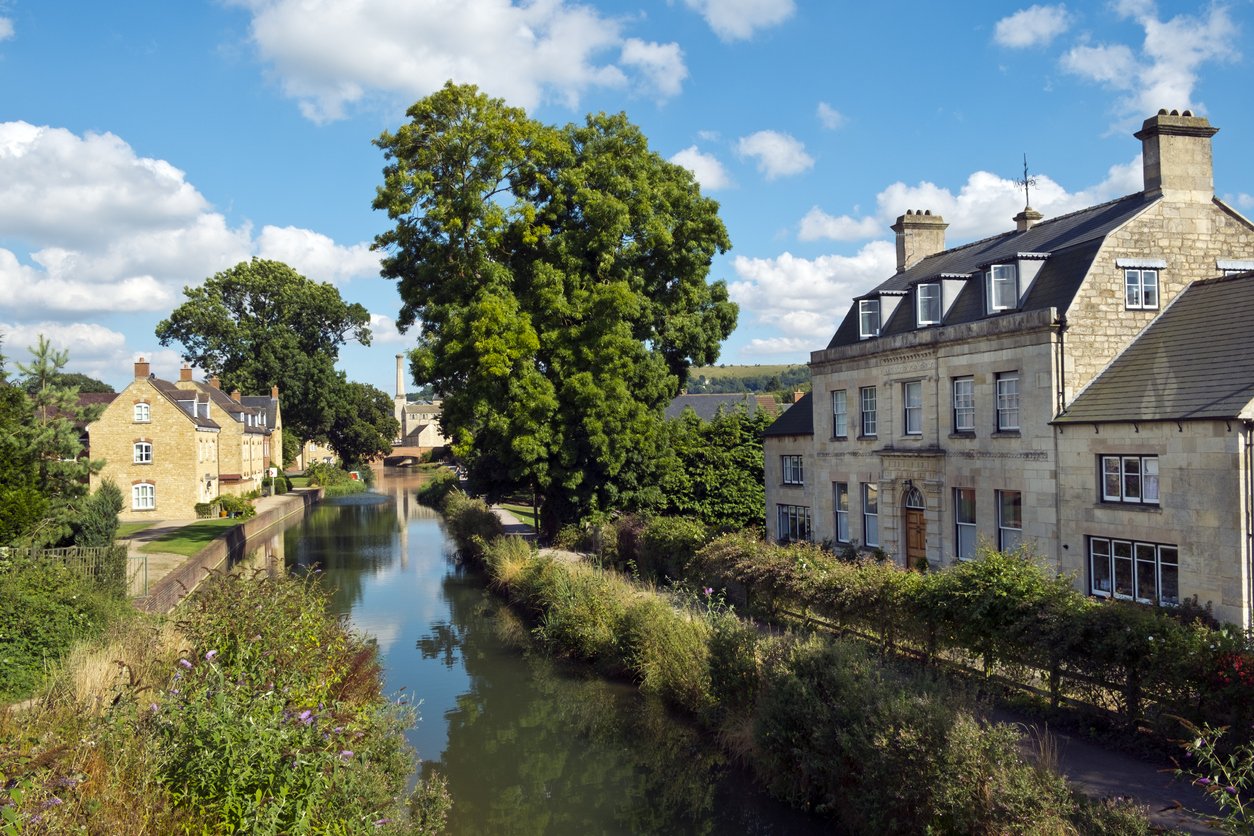 A scenic view of a river lined with greenery and historic stone houses under a bright blue sky with white clouds.