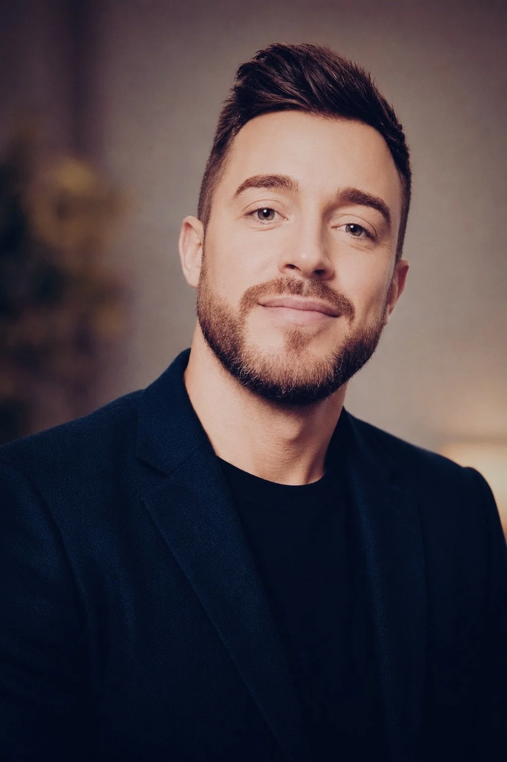 A young man with short dark hair, a beard, and blue eyes, smiling in a dark blazer and black shirt, with a blurred background.