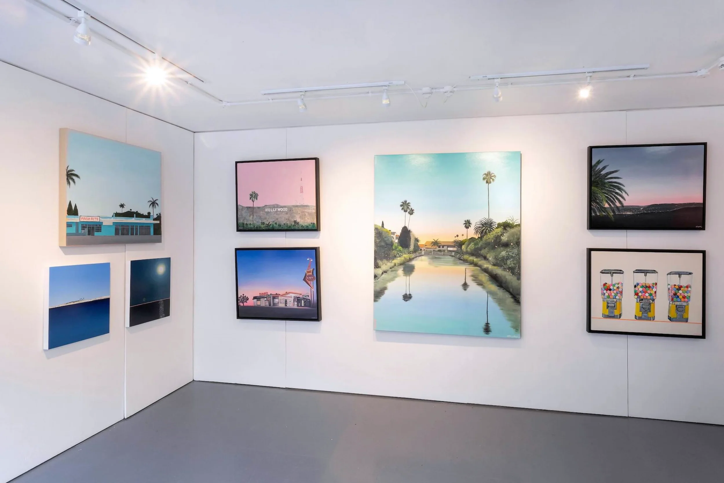 Gallery wall with paintings of Los Angeles landmarks, nature scenes, and a vintage gumball machine in a contemporary art gallery.