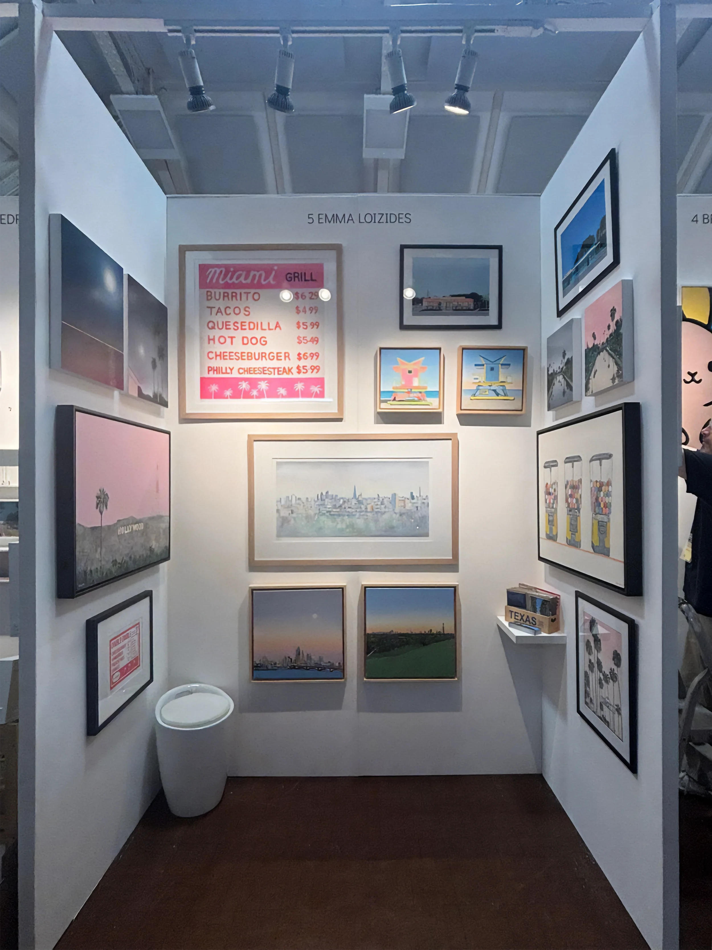 Art exhibit booth featuring framed paintings of cityscapes, landscapes, and pop art, a red menu board with food items and prices, and a small white trash can on a wooden floor.