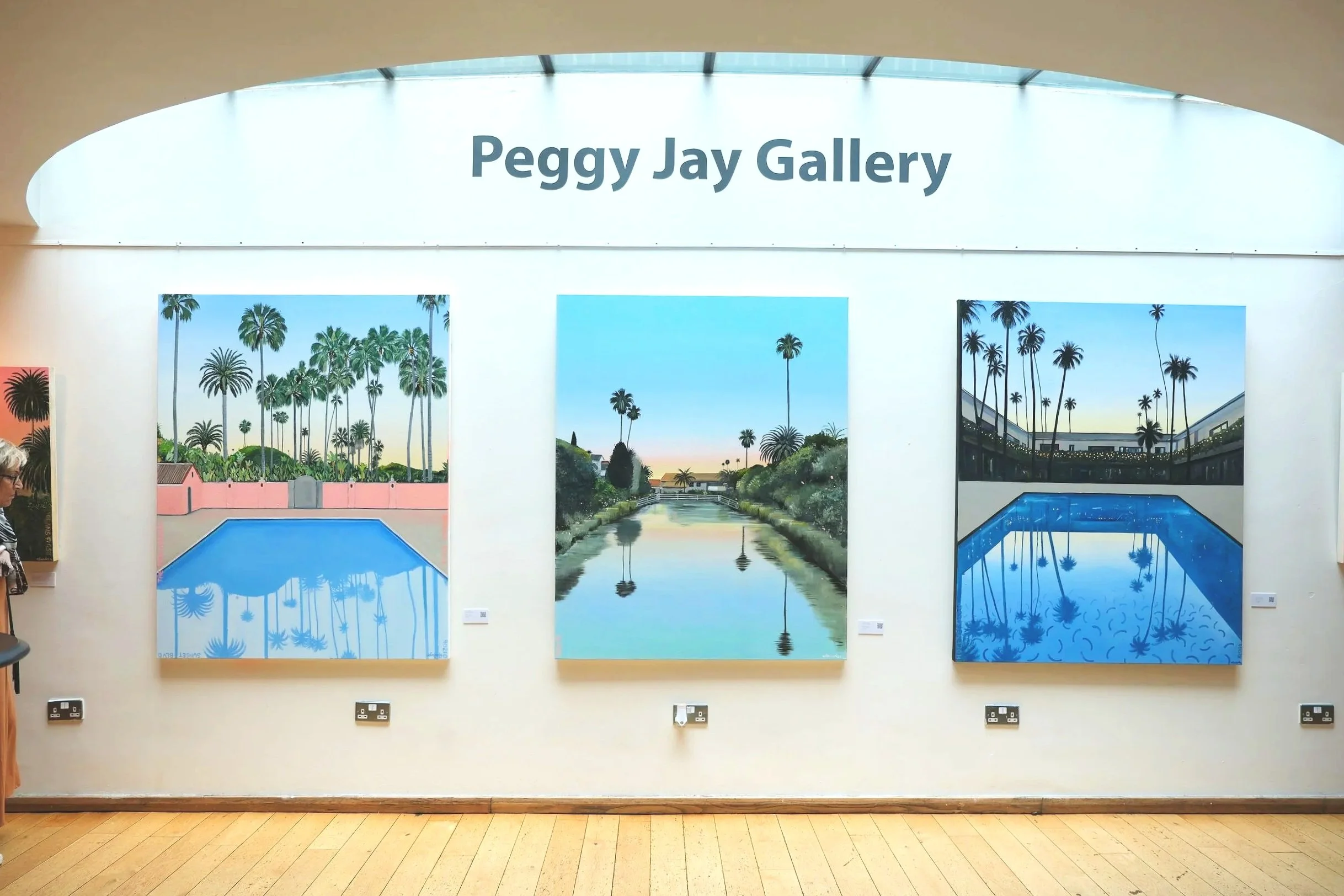Three landscape paintings of scenes with palm trees at Peggy Jay Gallery, depicting pools, waterways, and sunset sky.