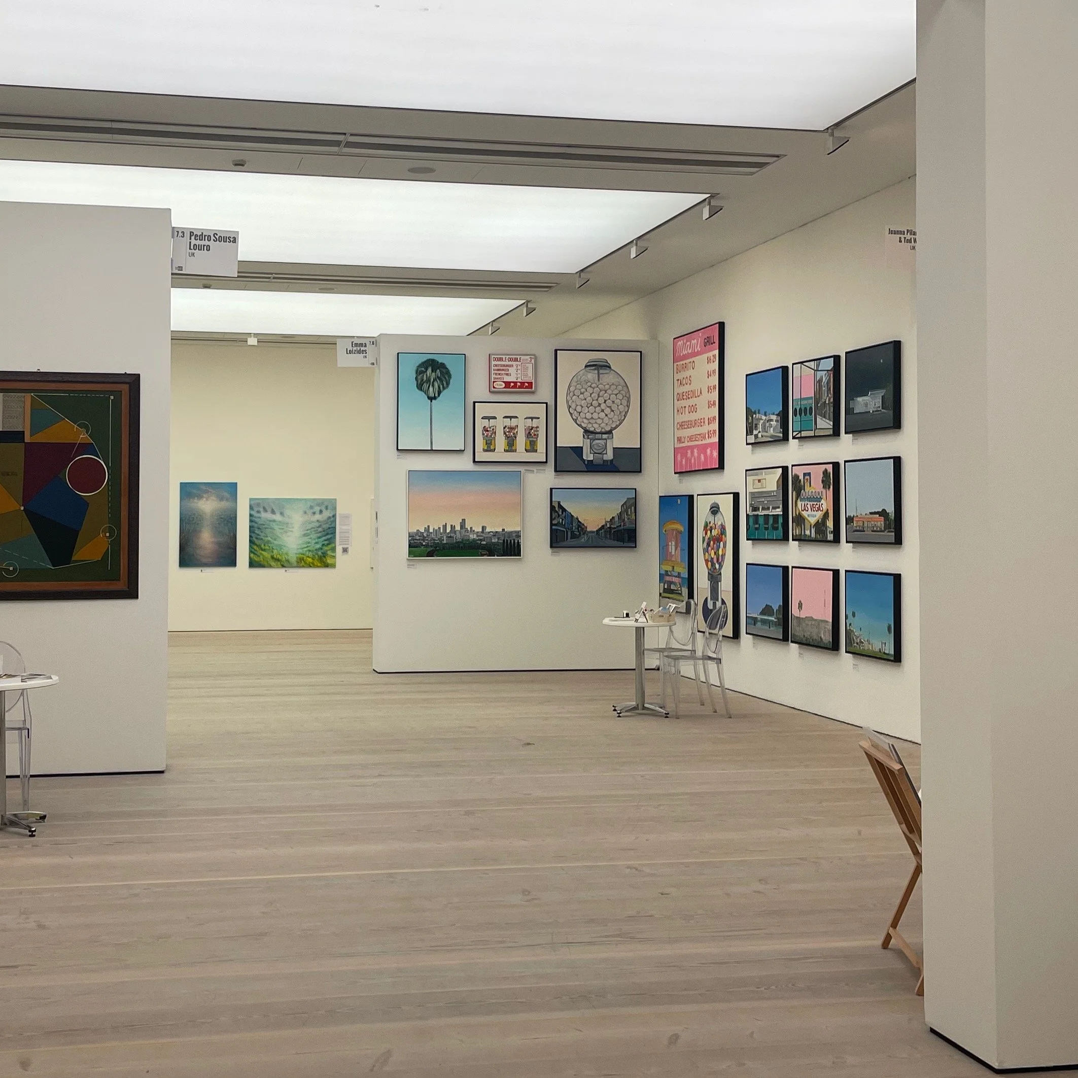Interior view of an art gallery with various framed paintings and photographs on white walls, featuring landscapes, cityscapes, and abstract art, with a light wood floor.