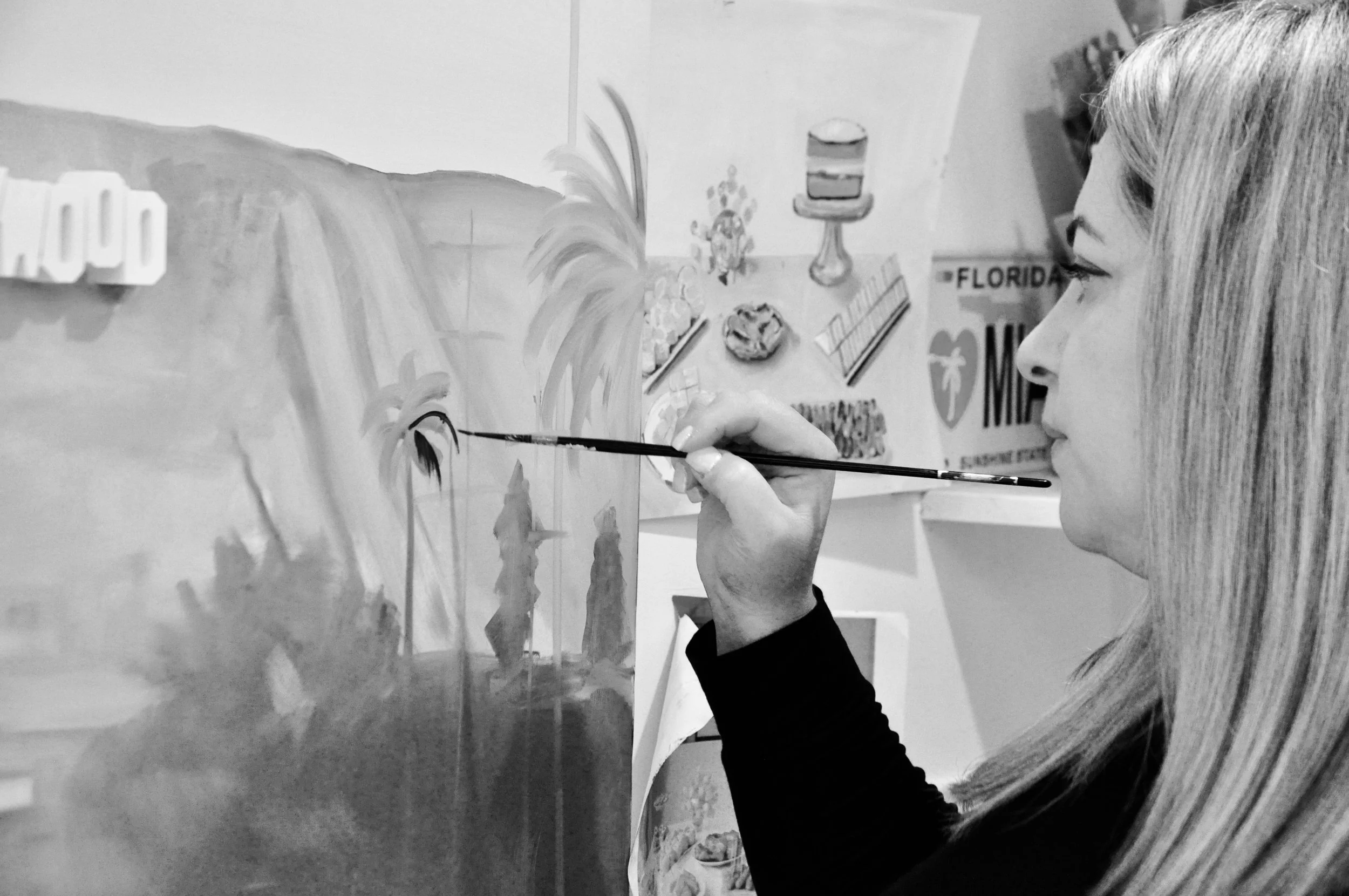Emma Loizides painting on a canvas. She is using a thin brush and has her right hand raised to work on the artwork, which features a landscape with palm trees and mountains.
