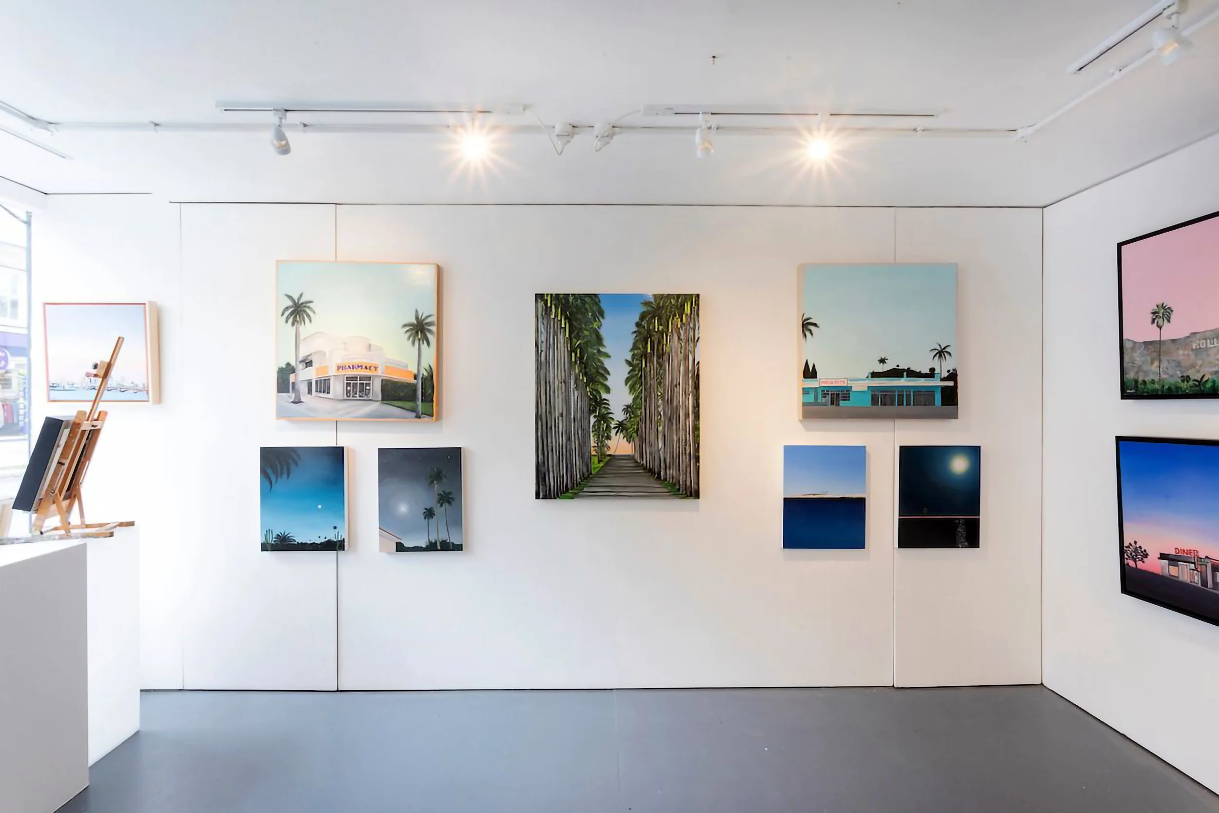 Gallery wall with various landscape paintings and photographs, featuring scenes of palm trees, buildings, and skies, in a brightly lit room.