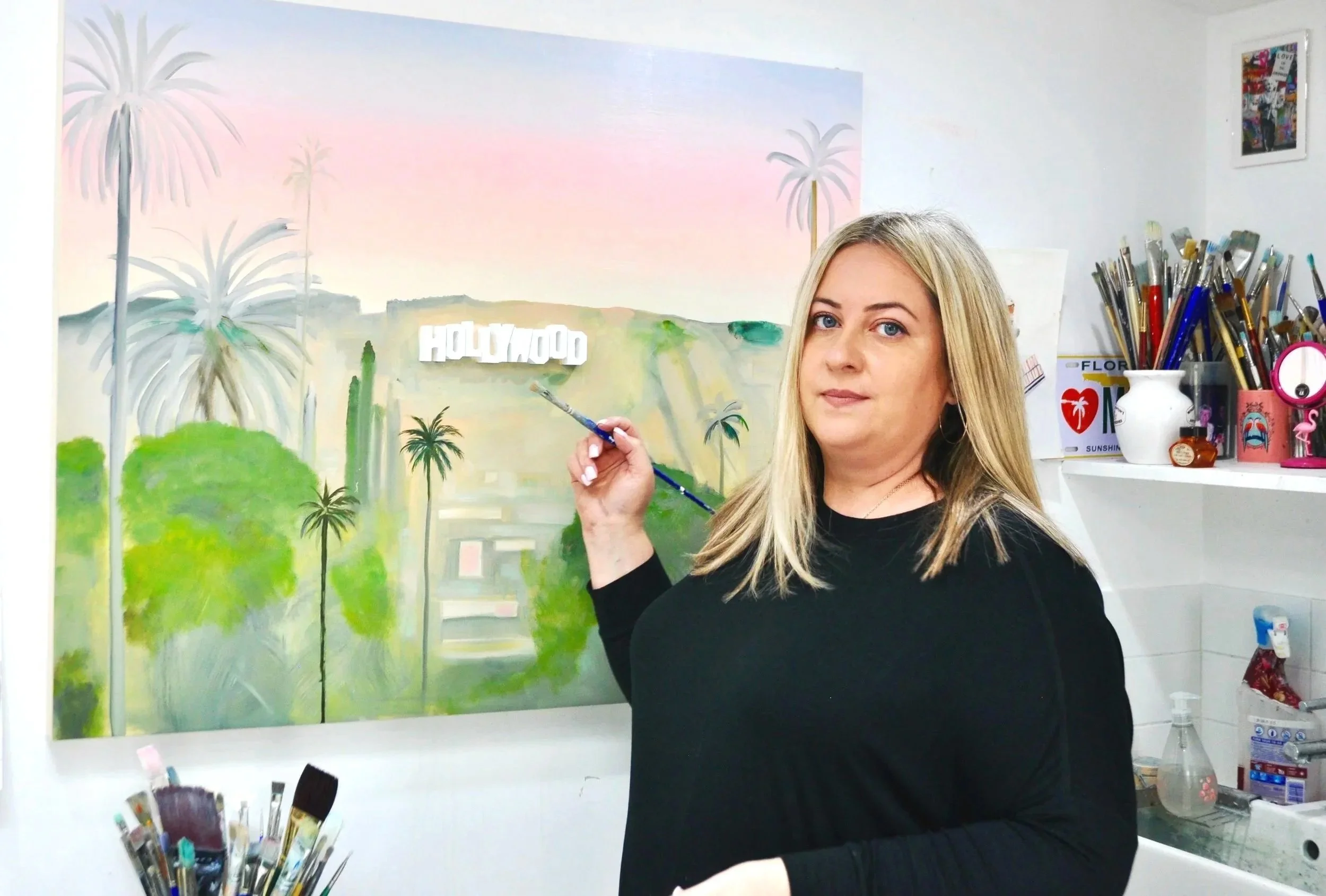 Emma Loizides holding a paintbrush in front of an oil painting of Hollywood sign and palm trees in a studio or art space.
