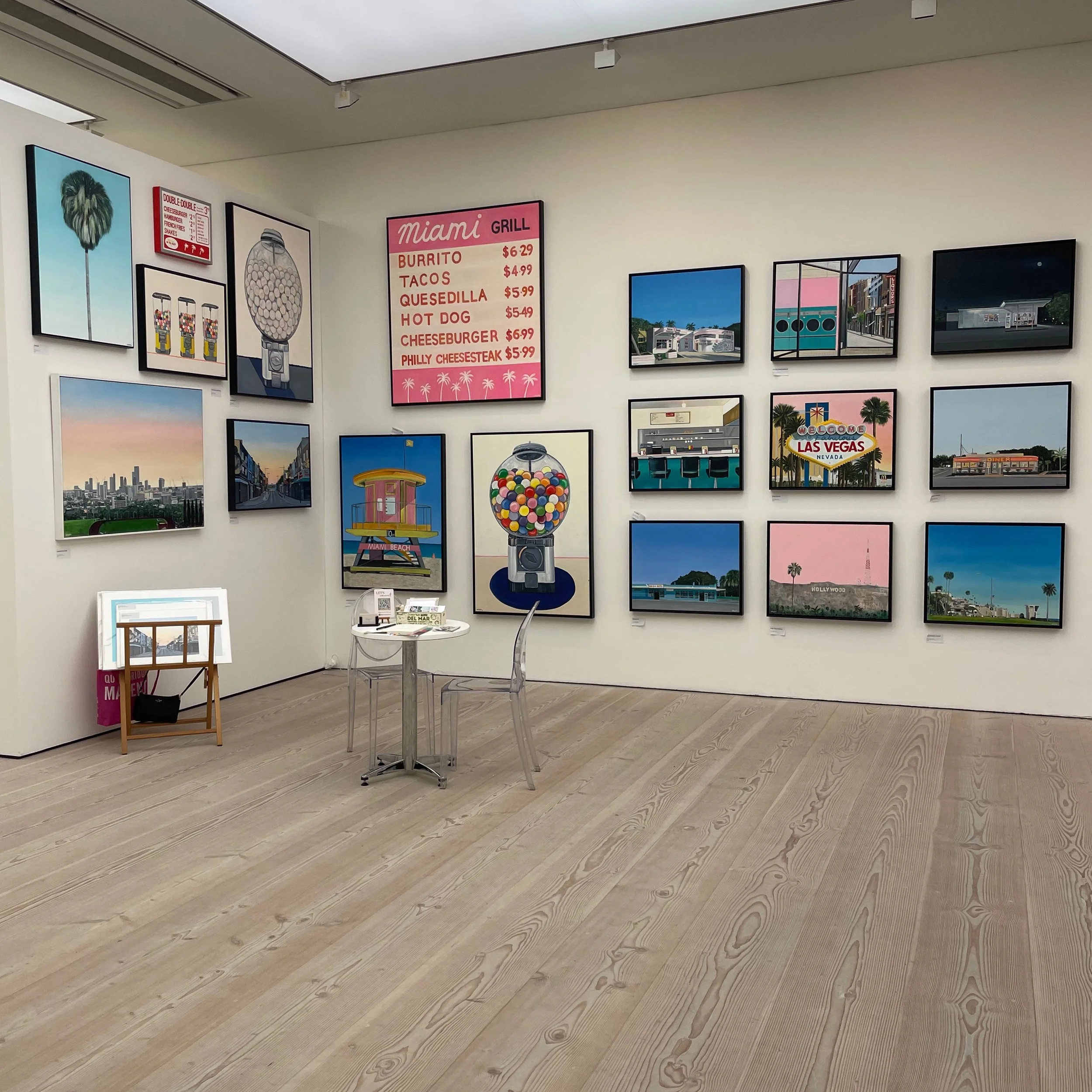 An art gallery with various framed paintings and photographs on the walls, including cityscapes, neon signs, and pop art images. There is a pink menu board listing food items and prices, a small table with chairs, and an easel with artwork on it. The gallery has light wood flooring and a neutral ceiling.