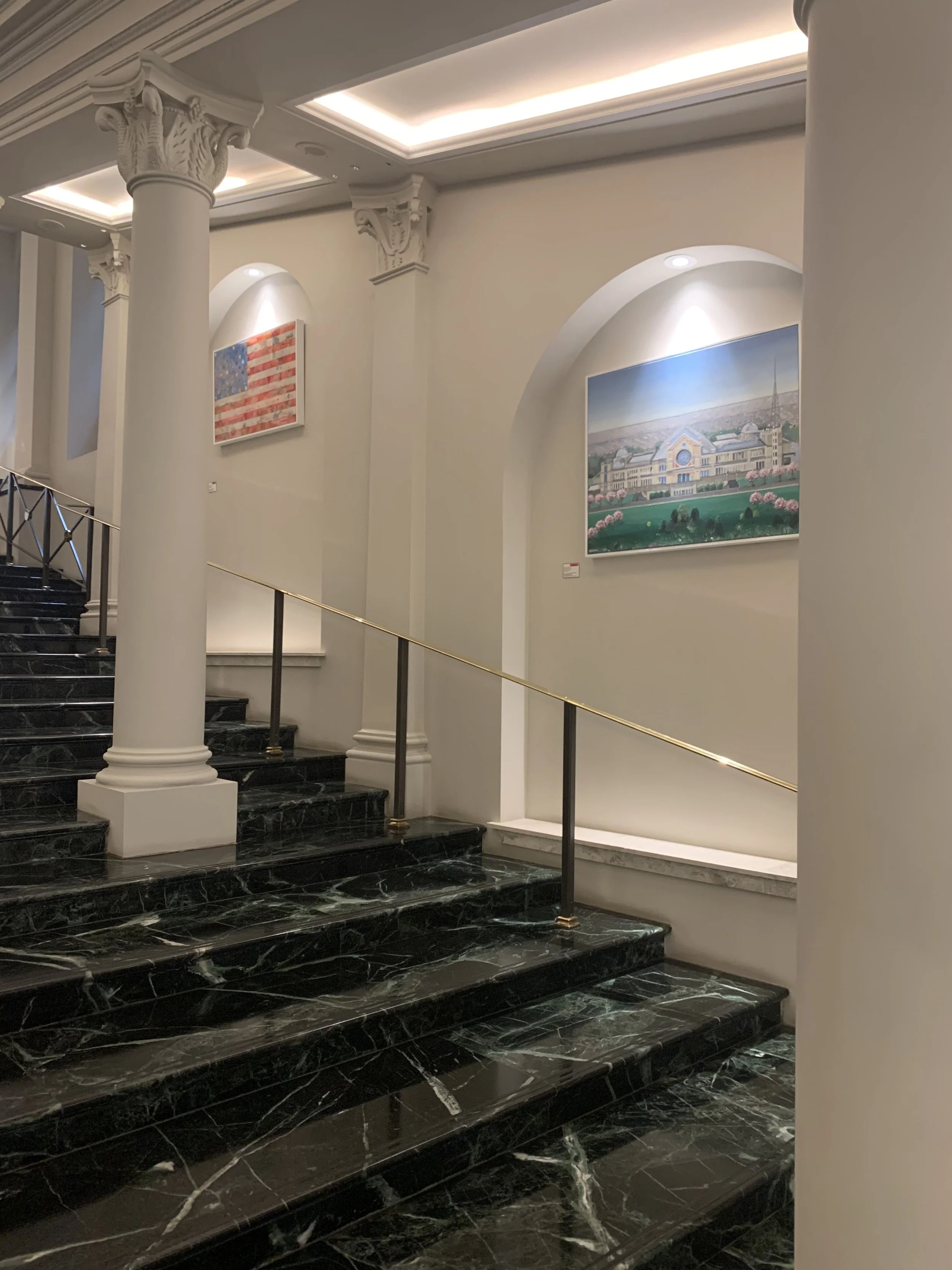 Interior view of a staircase with black marble steps, white columns, and framed artwork on the walls, including an American flag and a landscape painting.