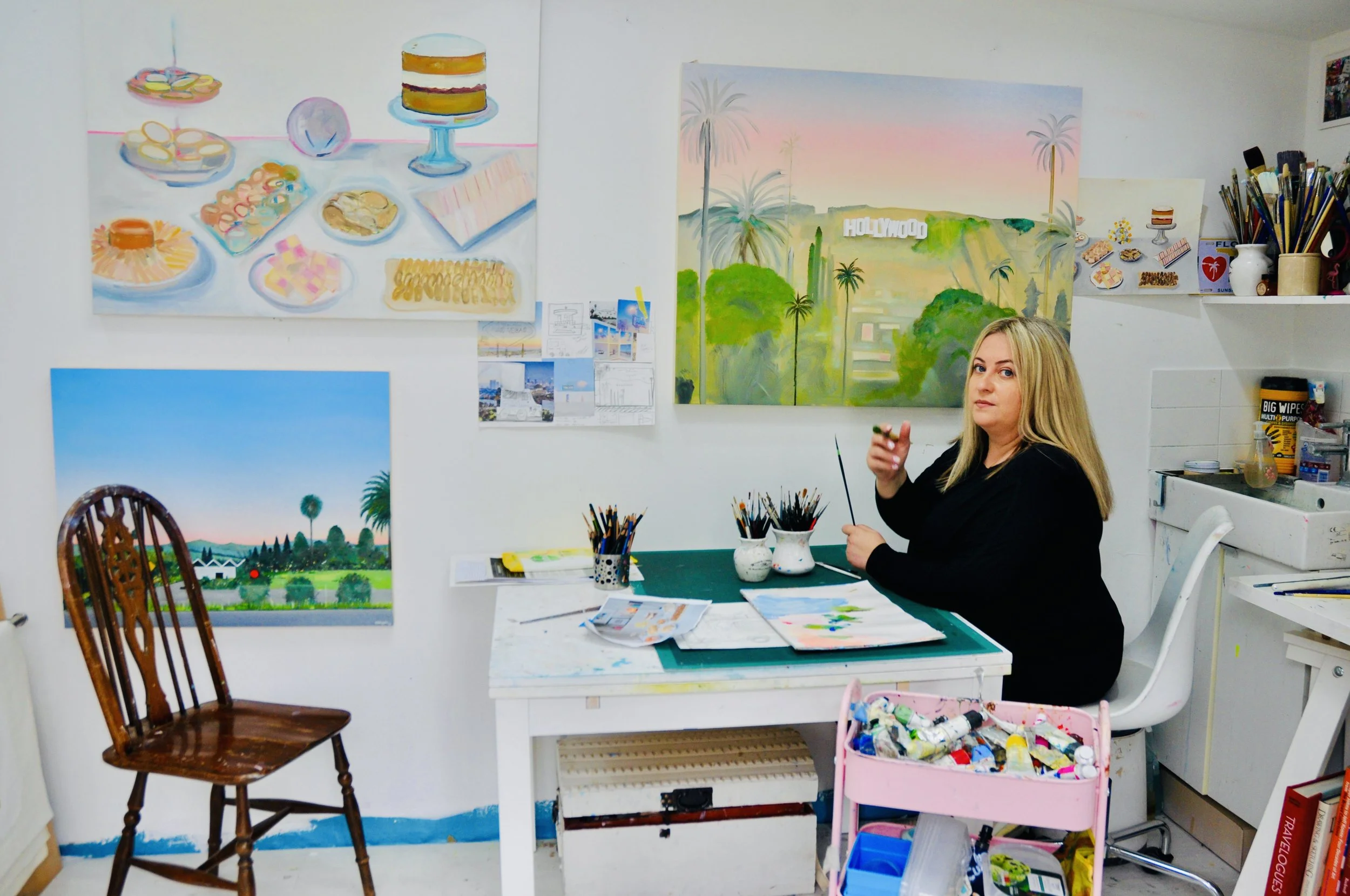 Emma Loizides sitting at an art studio table, holding a paintbrush, surrounded by paintings, art supplies, and a pink cart filled with paint tubes.