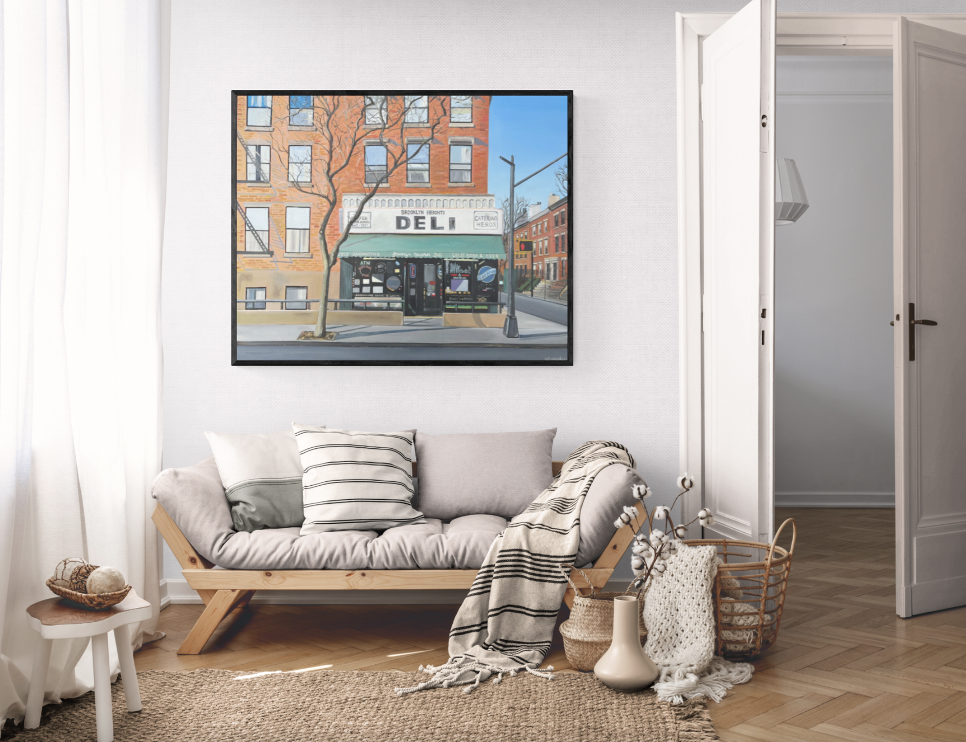 Living room with gray sofa, striped throw blanket, cushions, woven basket with cotton branches, ceramic vase, small side table with yarn balls, framed urban art on white wall, white curtains, wooden floor, open door to another room.