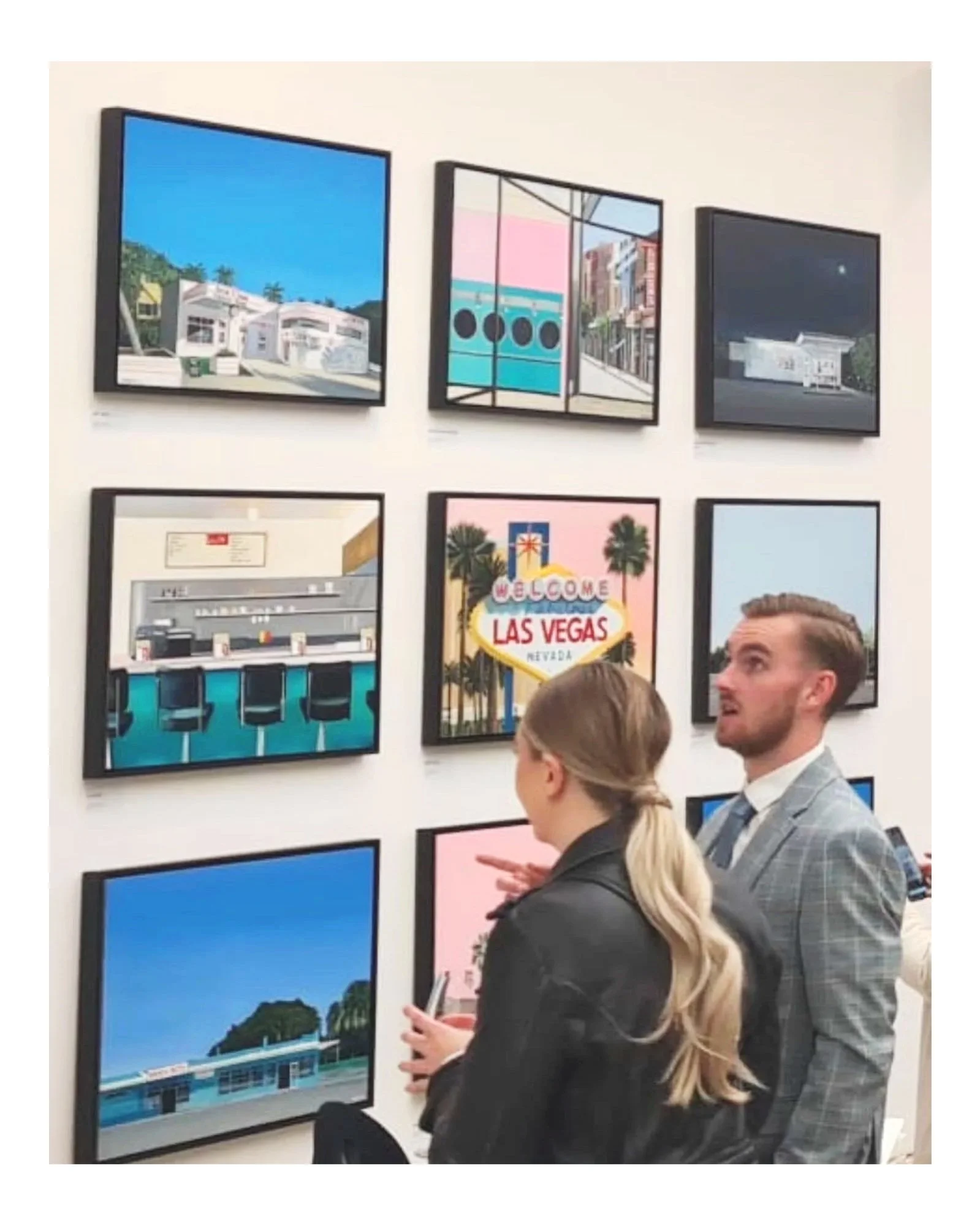 Two people, a man and a woman, viewing framed artwork on a gallery wall. The artwork depicts colorful scenes including a neon sign for Las Vegas.