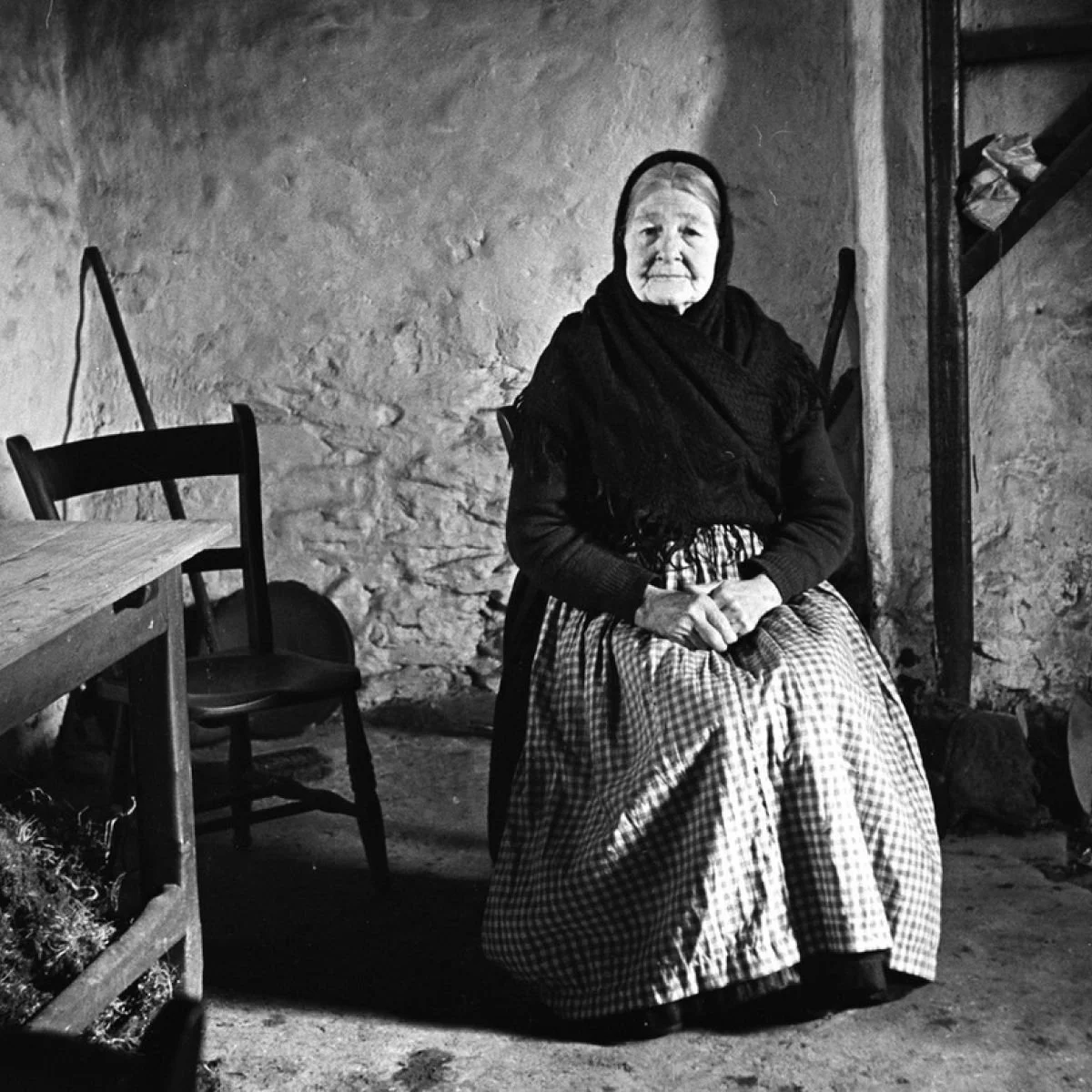 An elderly woman, Peig Sayers, wearing a headscarf, sitting on a chair in a rustic room with stone walls. She is wearing a checkered skirt and a dark top.