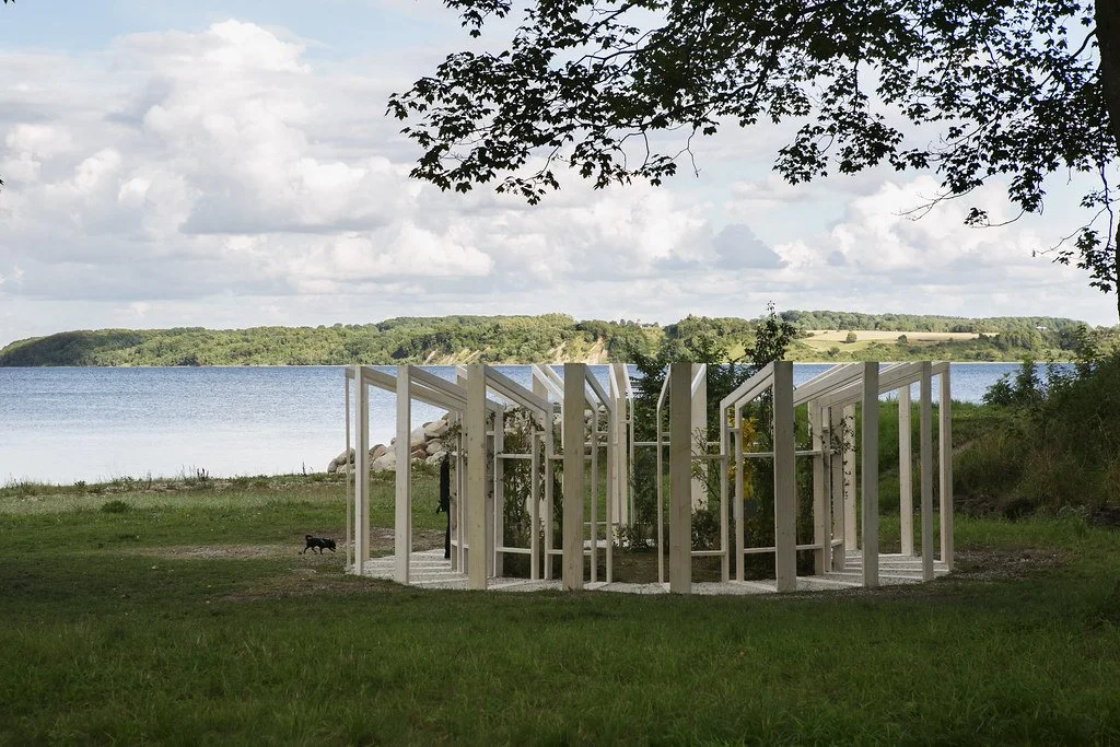 An outdoor scene featuring an architectural installation white wooden frame structure on a grassy area near a lake, with trees and hills in the background and a partly cloudy sky overhead.