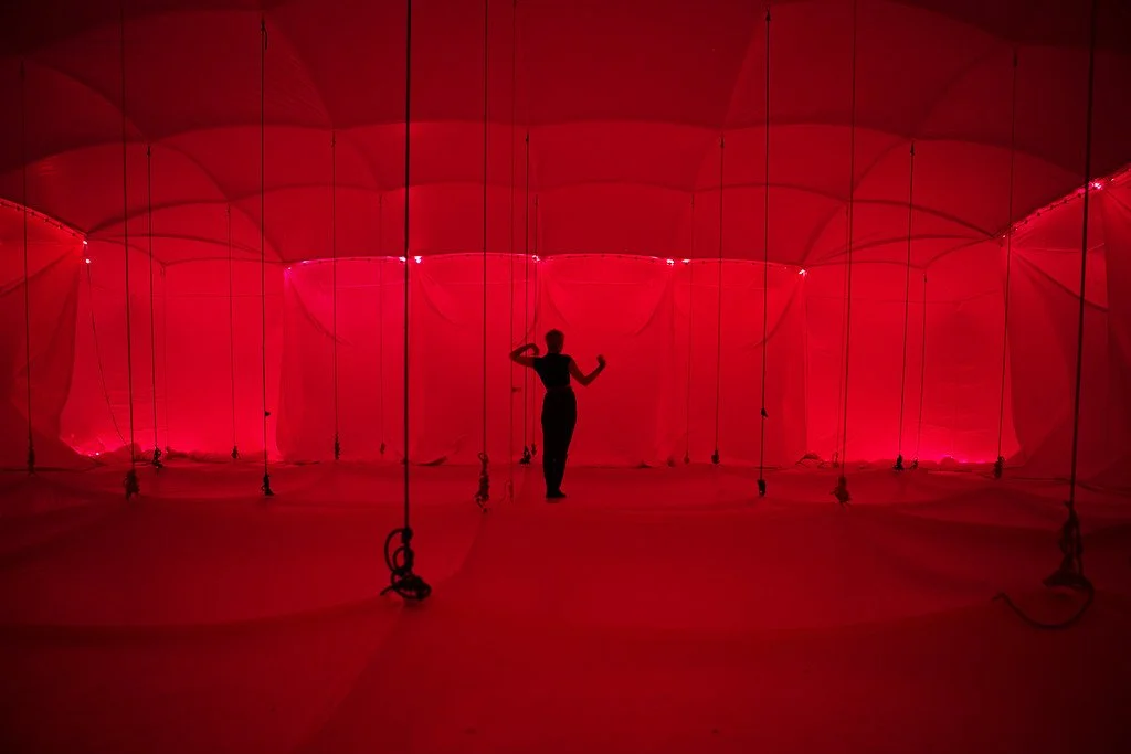 Person standing inside an architectural installation with red fabric walls, illuminated by red lighting, and held up by black ropes.