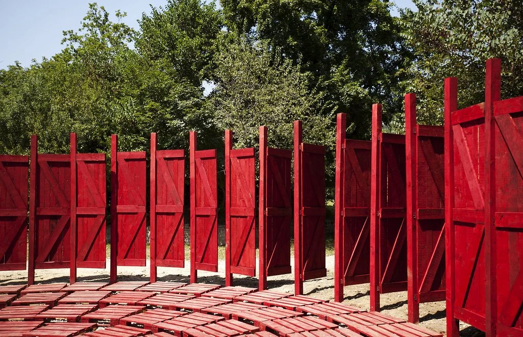 Red wooden architectural installation outdoors with green trees in the background.