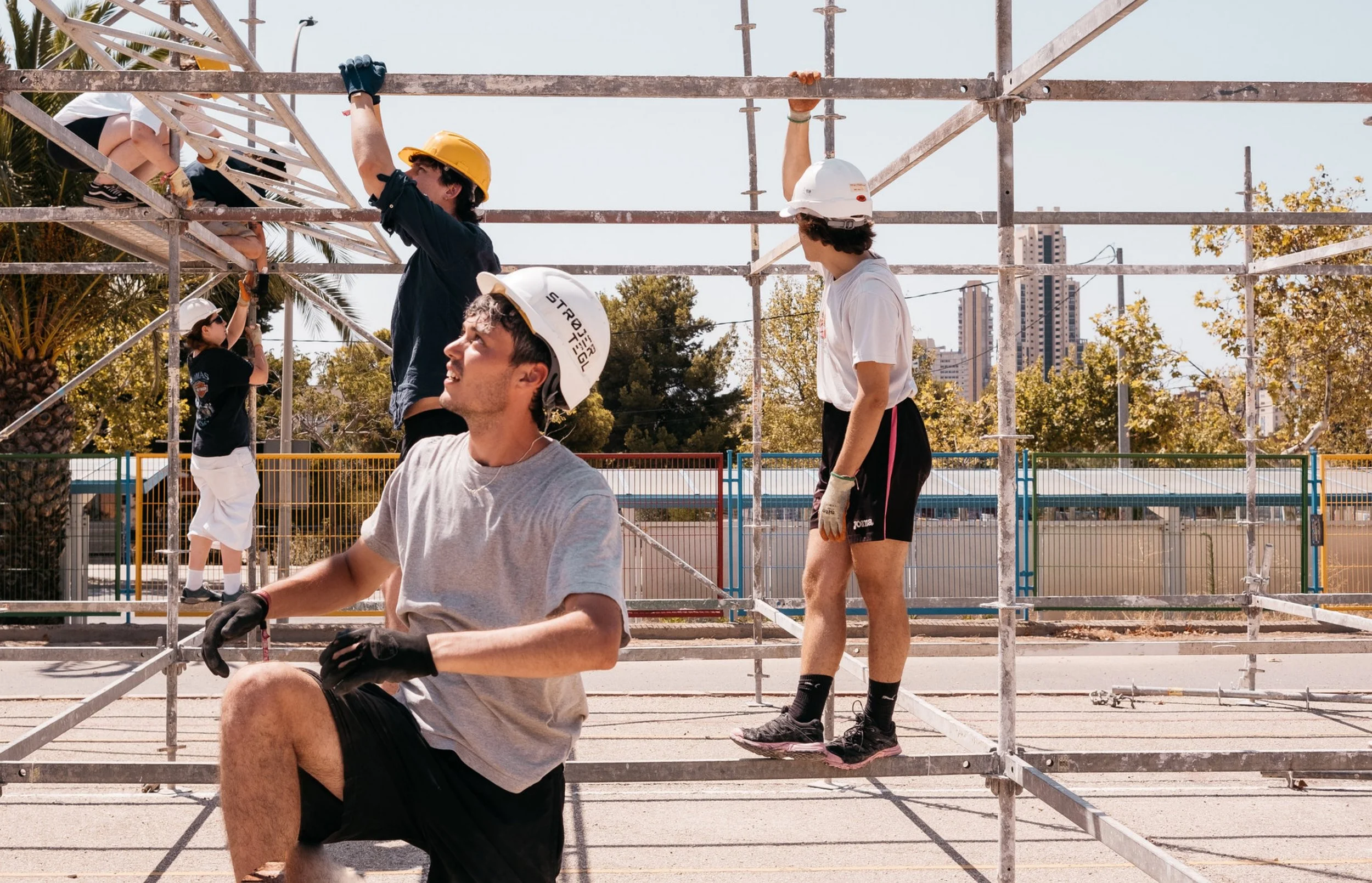 People installing scaffolding outdoors on a sunny day, some wearing helmets and gloves, with trees and city buildings in the background.