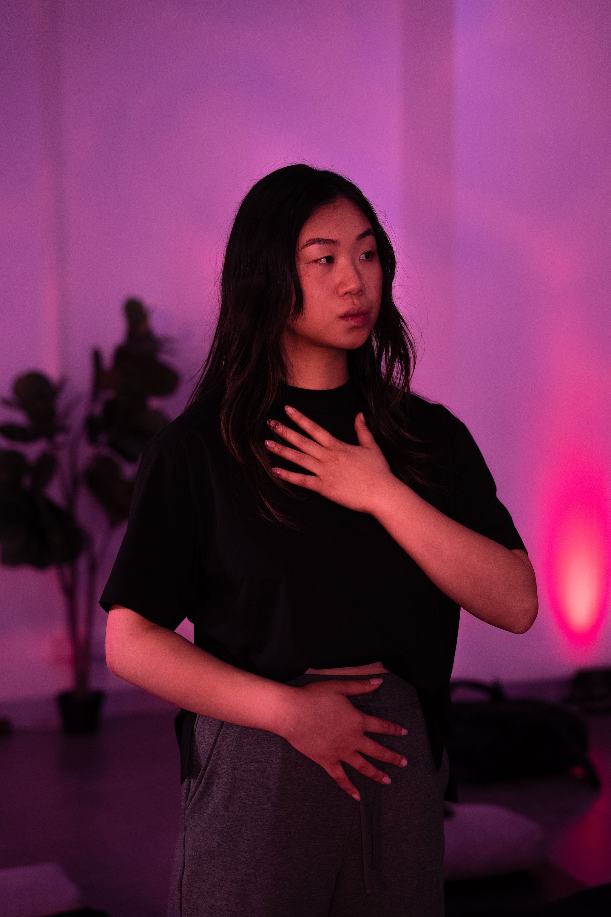 Liz Ho demonstrating a breath awareness exercise with one hand on her chest and one on her stomach during a breathwork session under purple lighting.