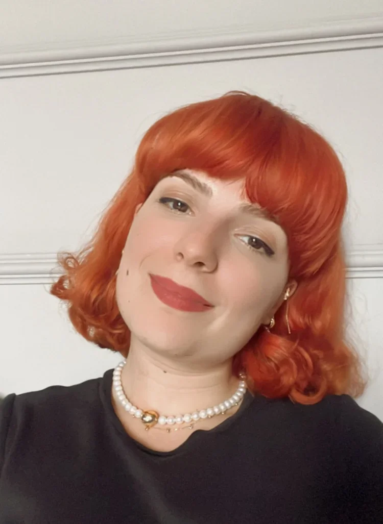 A woman with bright red, curly hair and fair skin, wearing a black top and a pearl necklace, smiling in front of a white wall.