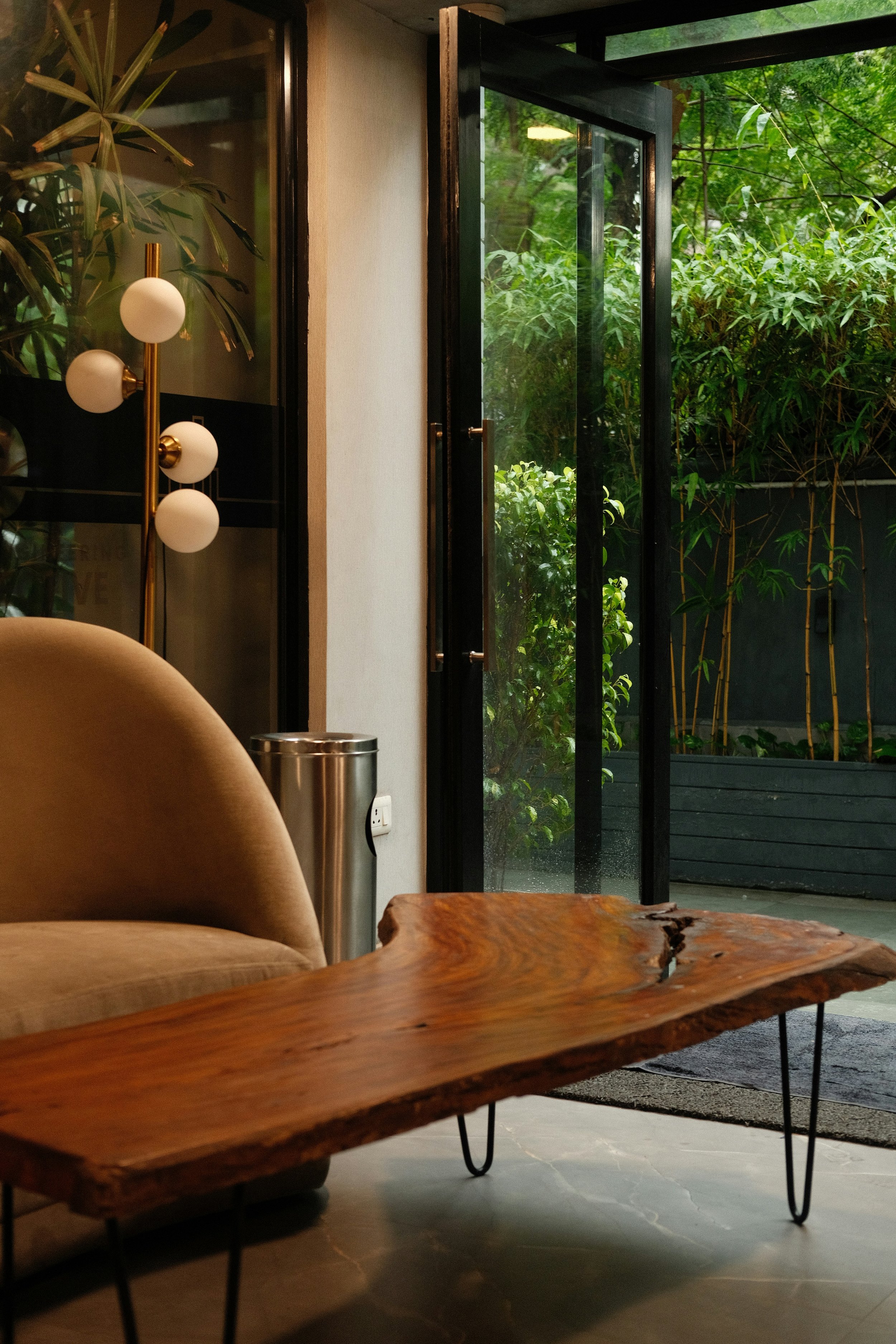 Interior of a room with a beige sofa, a wooden coffee table, a tall lamp with white globes, and an open glass door leading outside to greenery with bamboo plants.