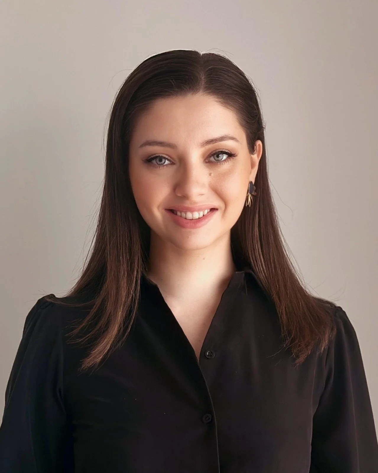 A woman with long brown hair, wearing a black blouse, smiling slightly, in front of a plain beige background.