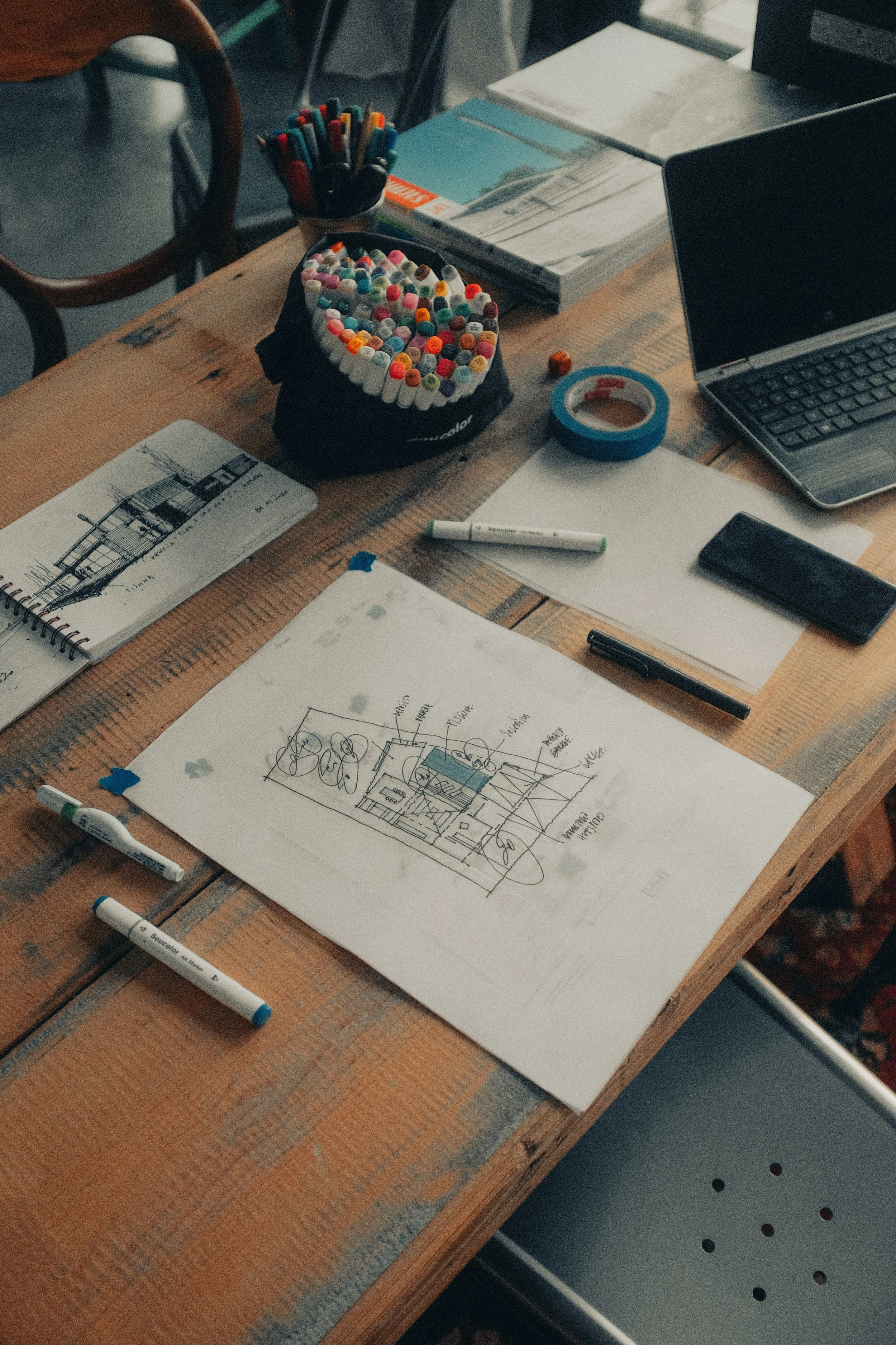 A wooden table with architectural sketches, markers, a book, a laptop, a phone, art supplies, and tape on it.