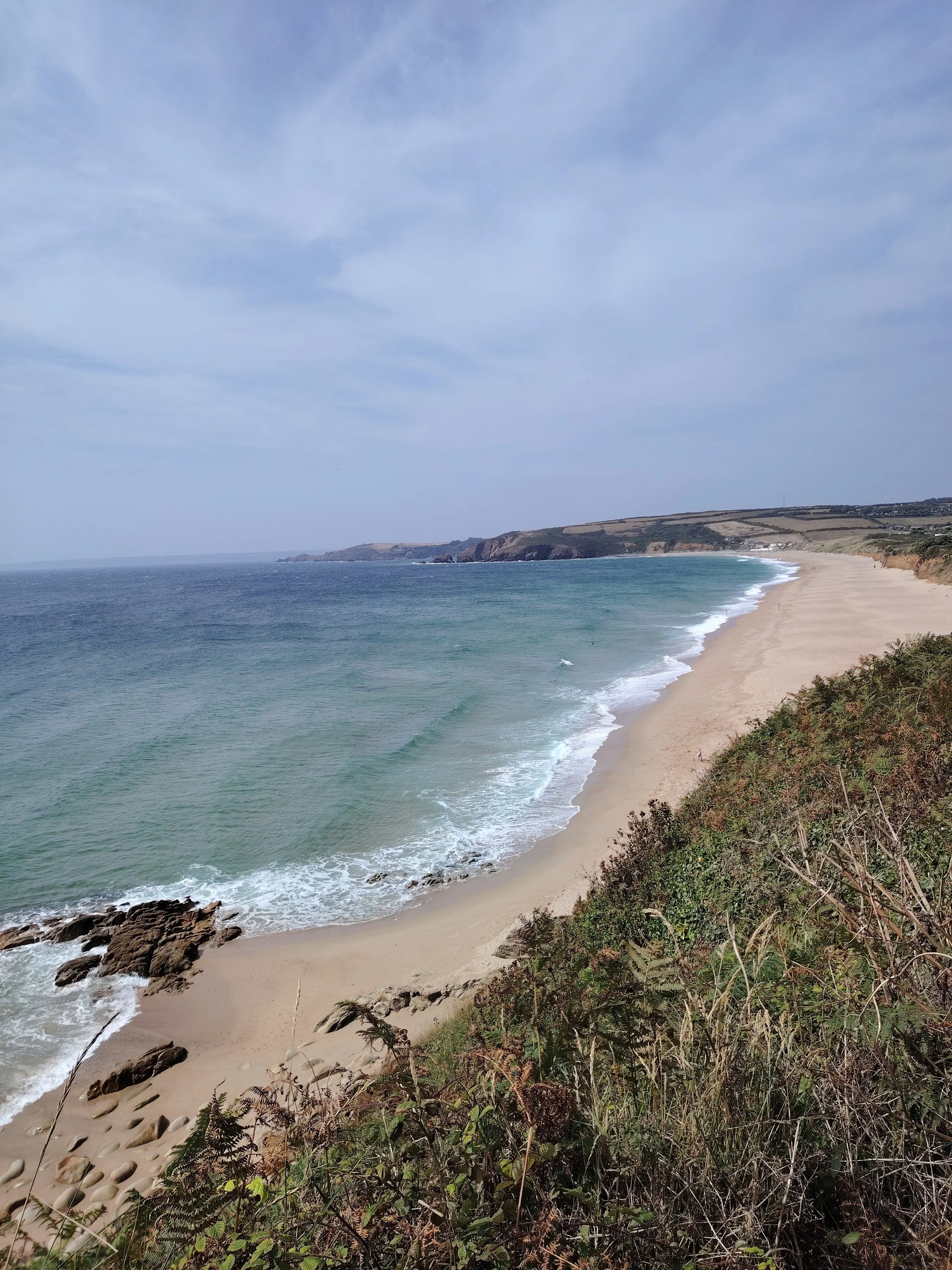 Beach with sandy shore, rocks, green vegetation, and the ocean under partly cloudy sky, with cliffs in the distance.